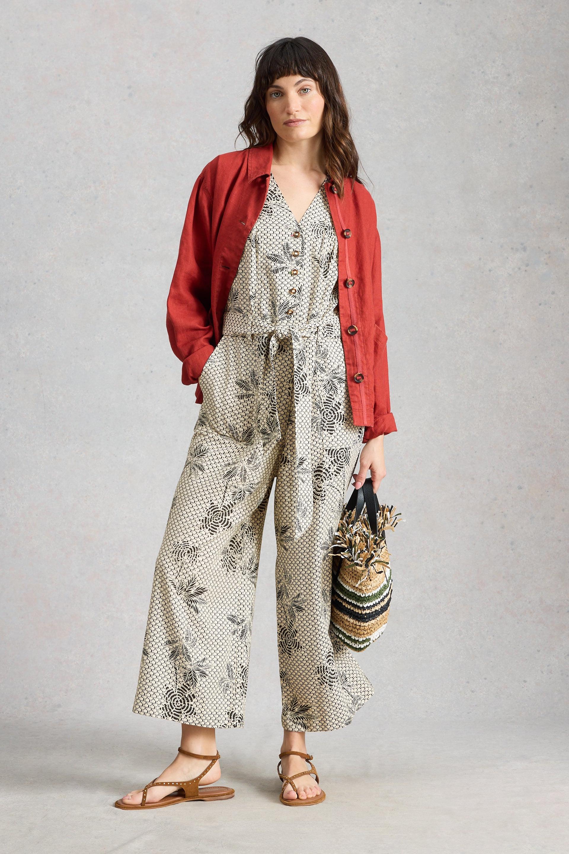 Woman wearing a red jacket, floral jumpsuit and holding a bag against a gray background