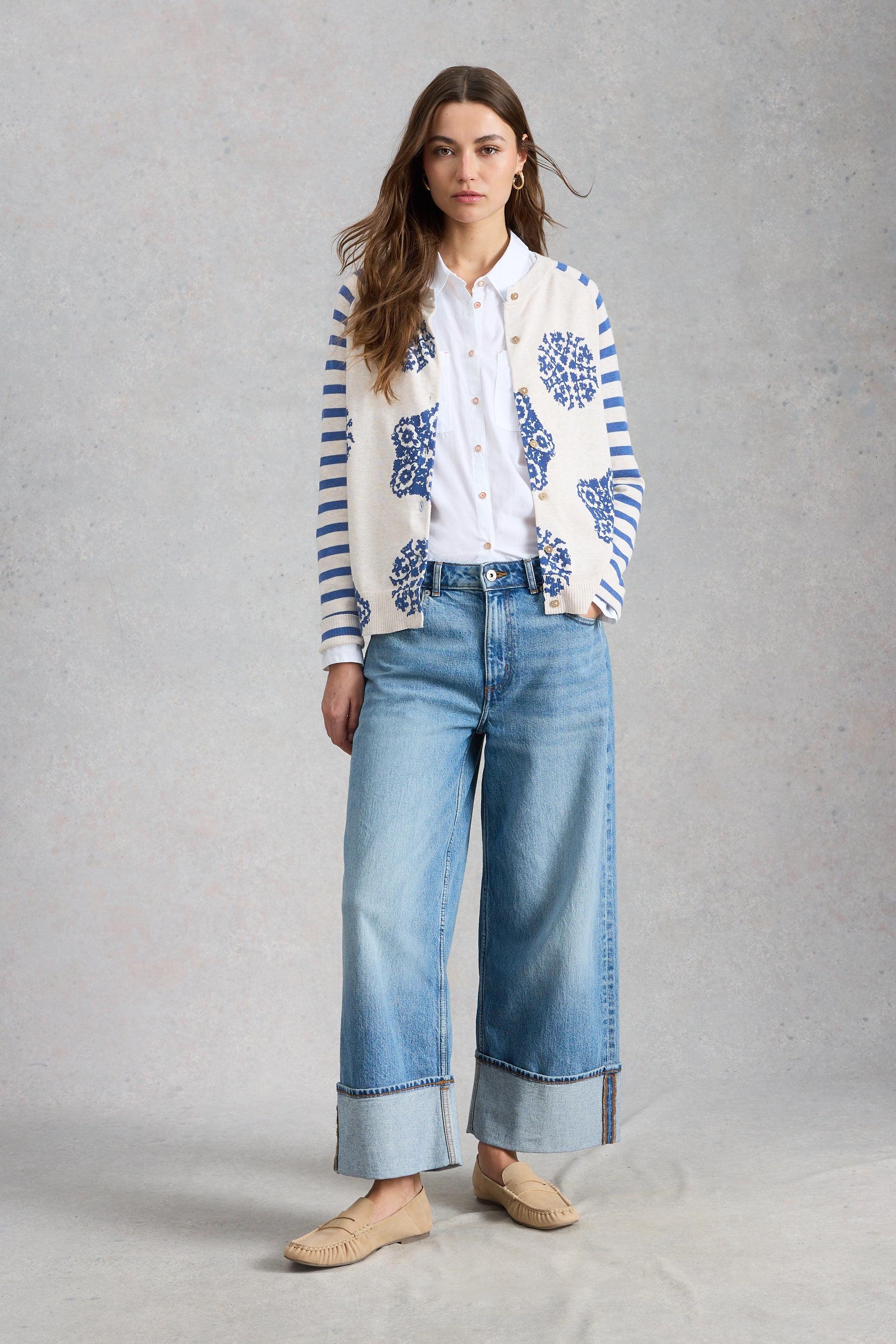 Woman wearing a patterned White Stuff cardigan, white blouse, blue jeans, and beige shoes on a gray background