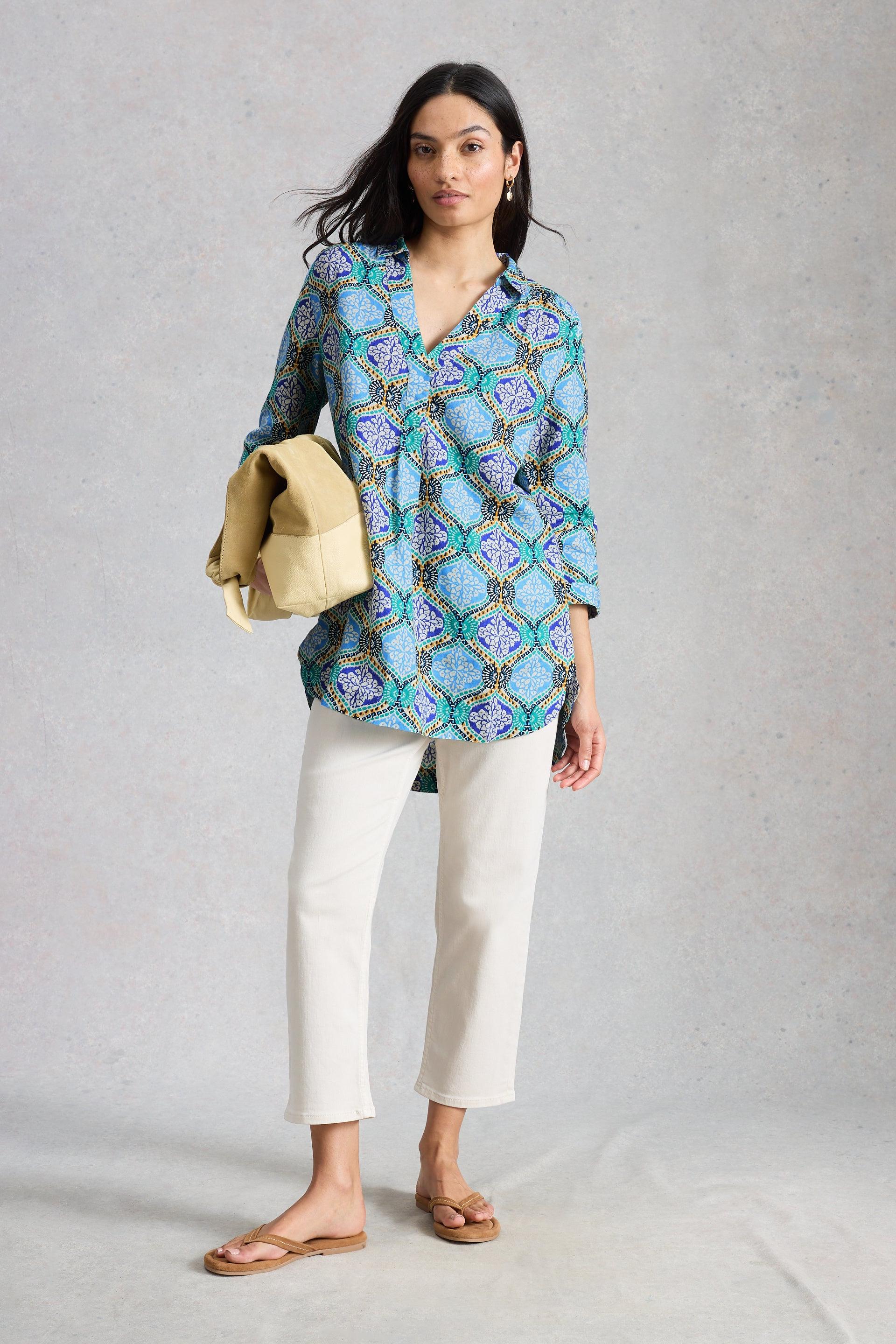 Woman wearing a blue patterned tunic and white pants against a gray background