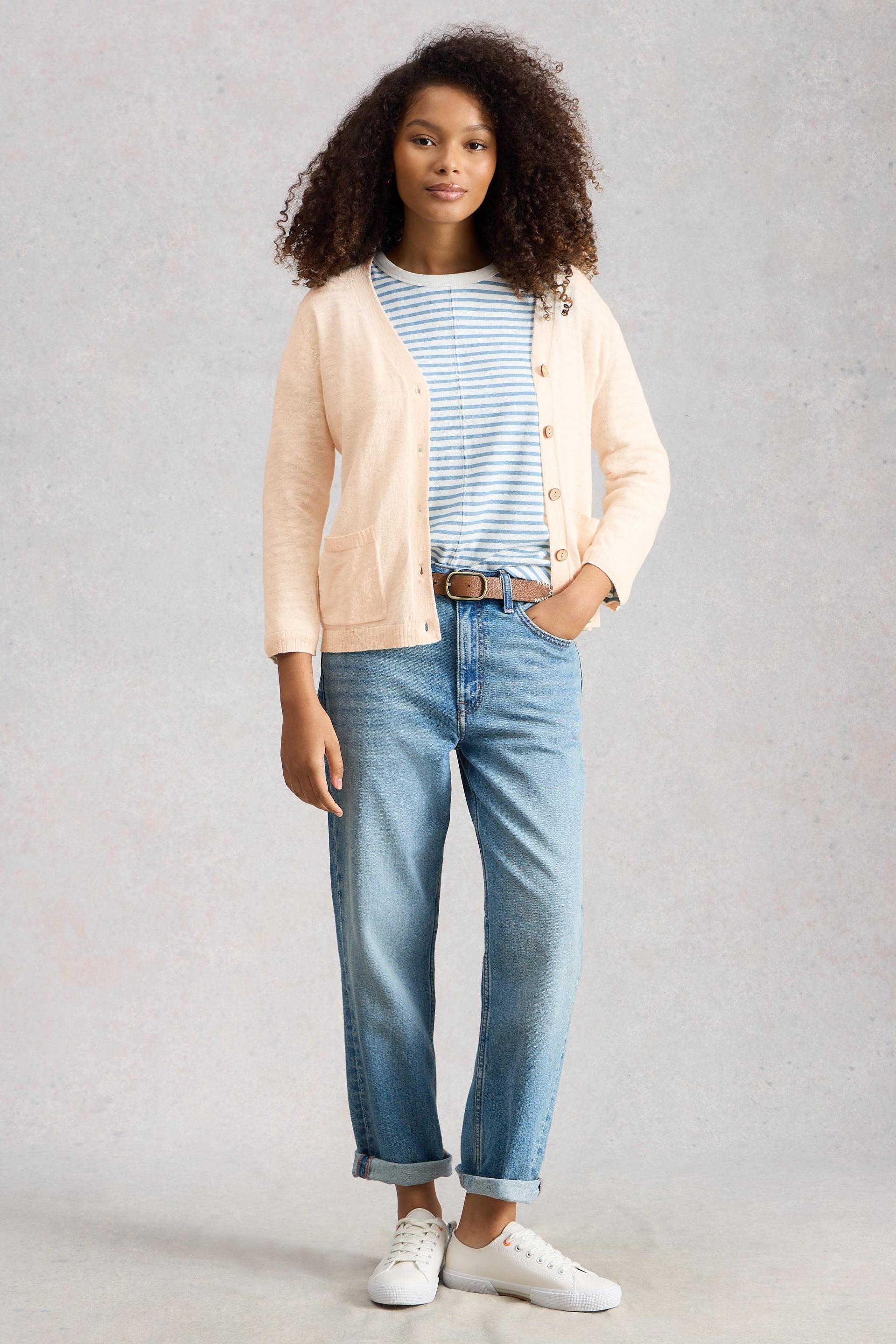 Person wearing a cream organic cotton cardigan, striped shirt, blue jeans, and white sneakers on a plain background