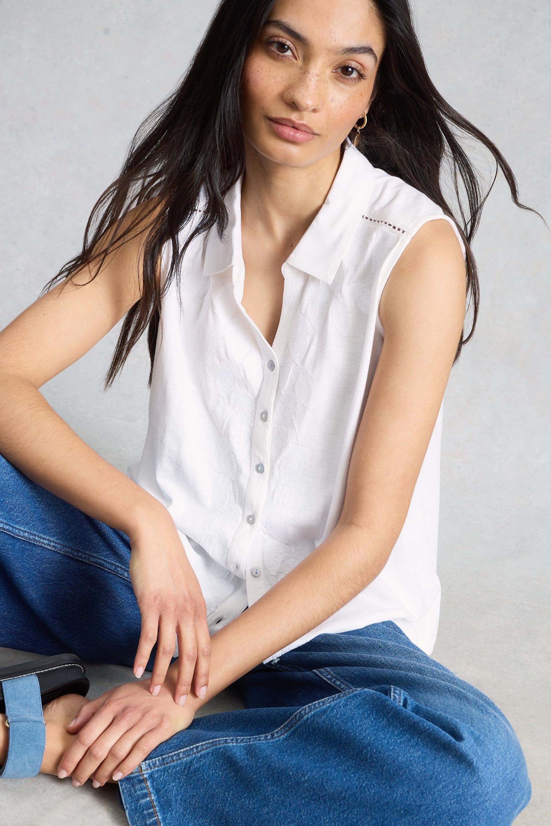 Woman wearing a white sleeveless shirt and blue jeans sitting on a gray background
