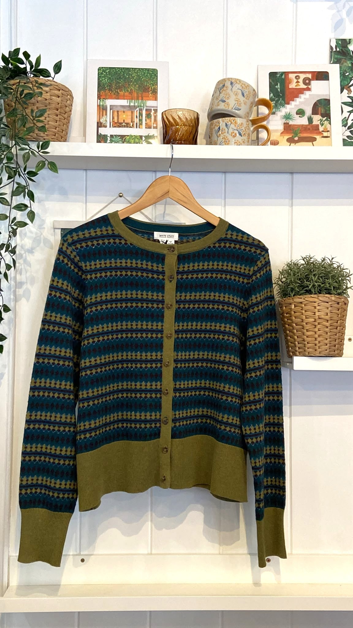 A patterned White Stuff fairisle button-down cardigan hanging on a wooden hanger against a white wall.
