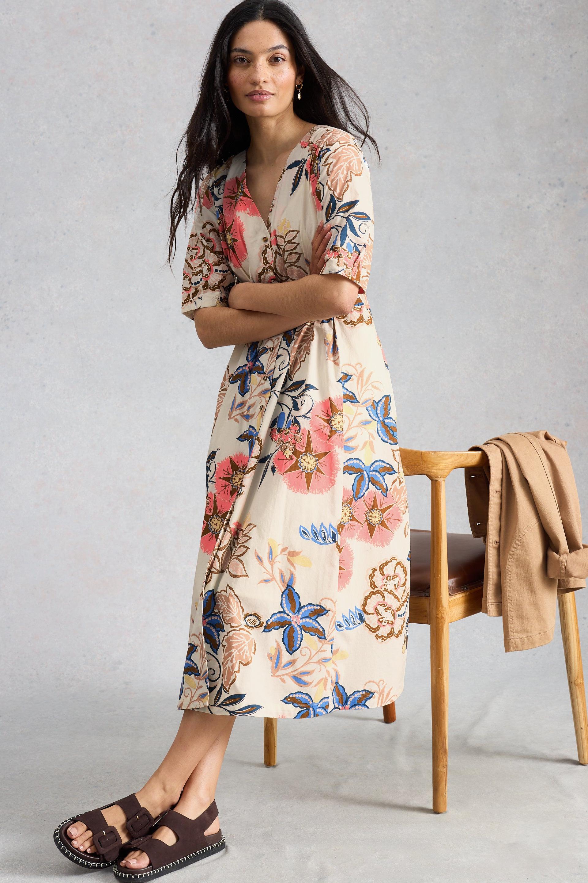 Woman wearing a floral organic cotton dress standing next to a wooden chair with a beige jacket on it.