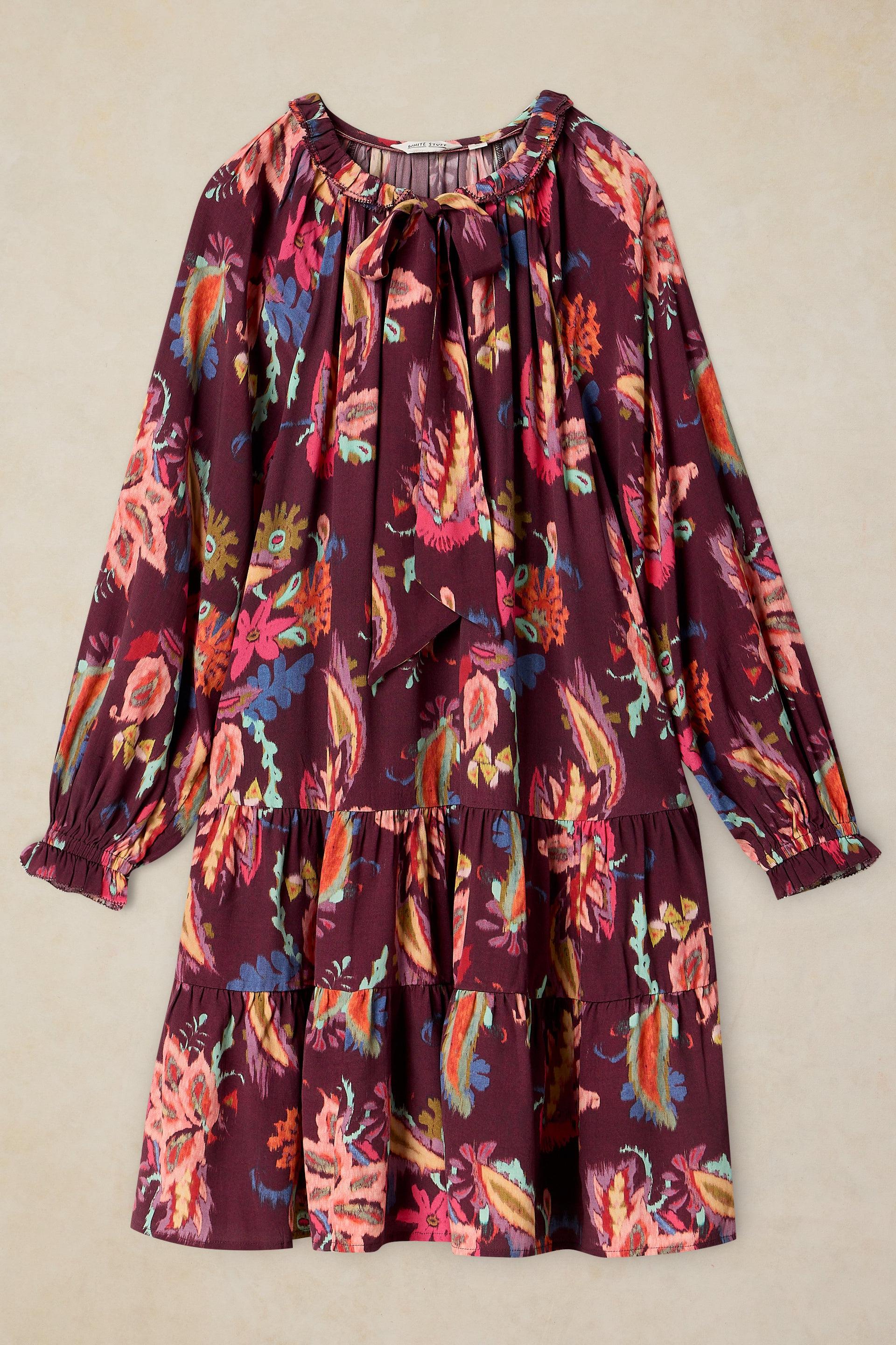 A long-sleeve tiered dress with a smocked bodice and a colorful floral print on a deep burgundy background, displayed flat against a neutral beige backdrop.