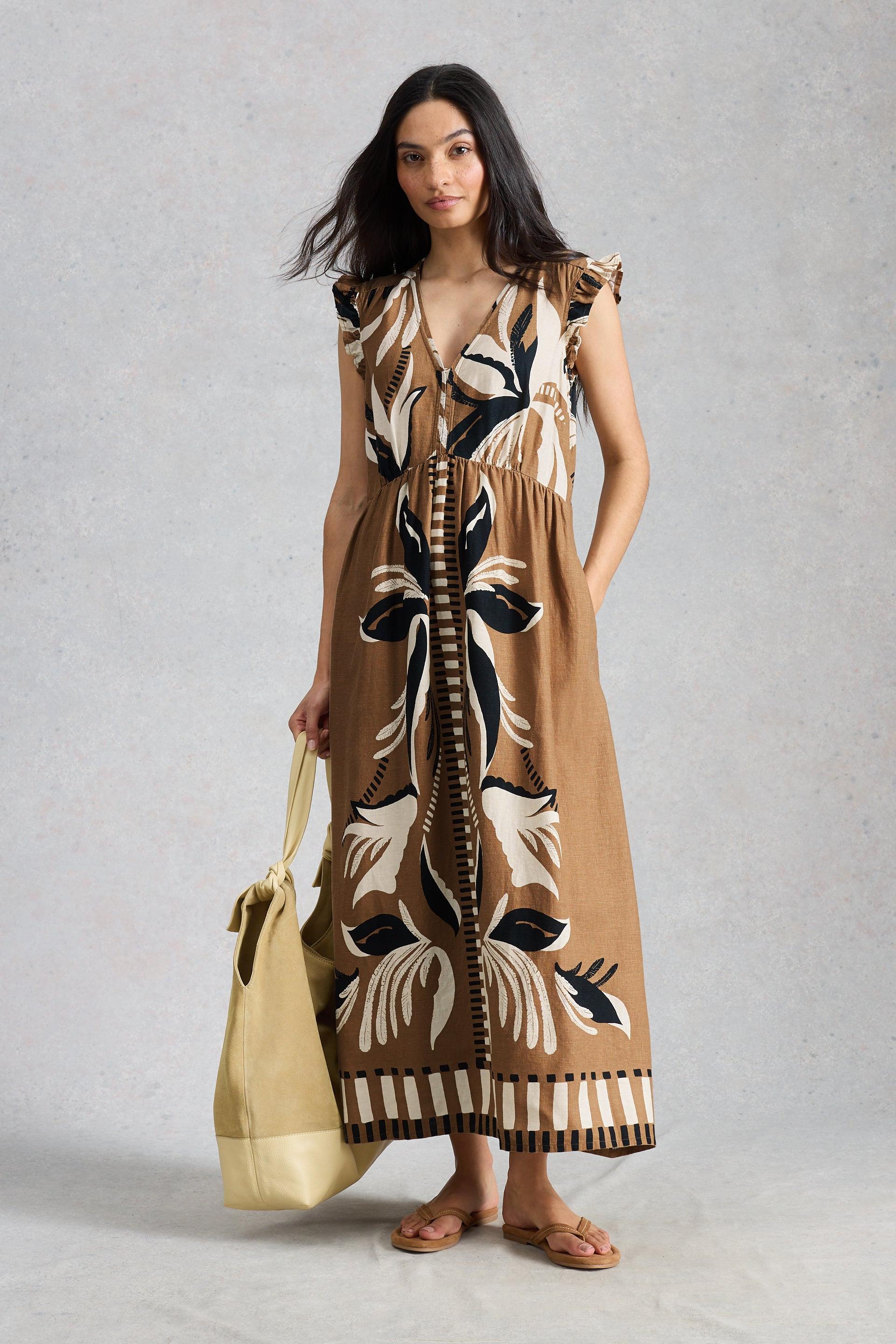 Woman wearing a long, patterned dress holding a beige handbag against a plain background