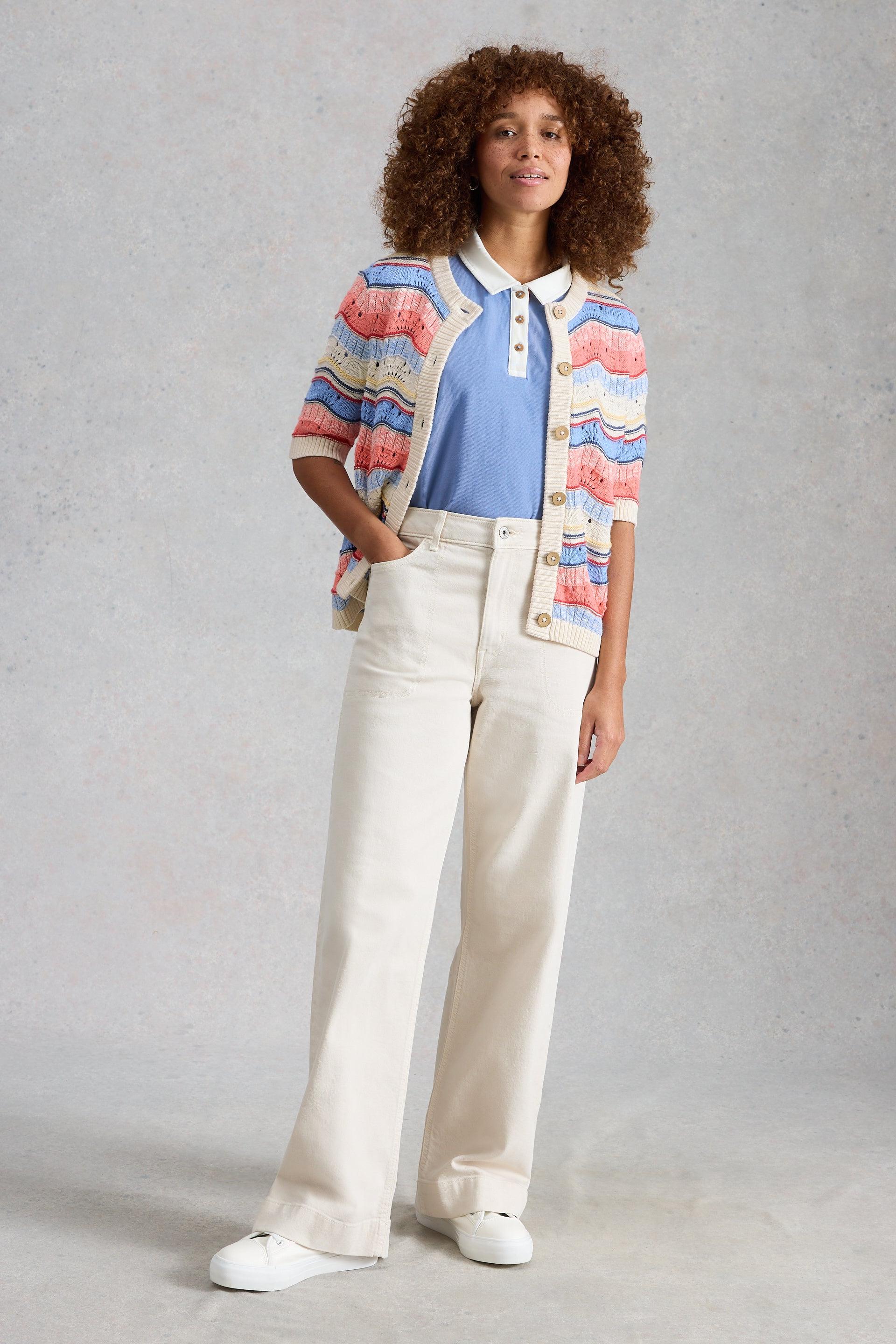Woman wearing a colourful striped cardigan, blue shirt, white pants, and white shoes on a gray background