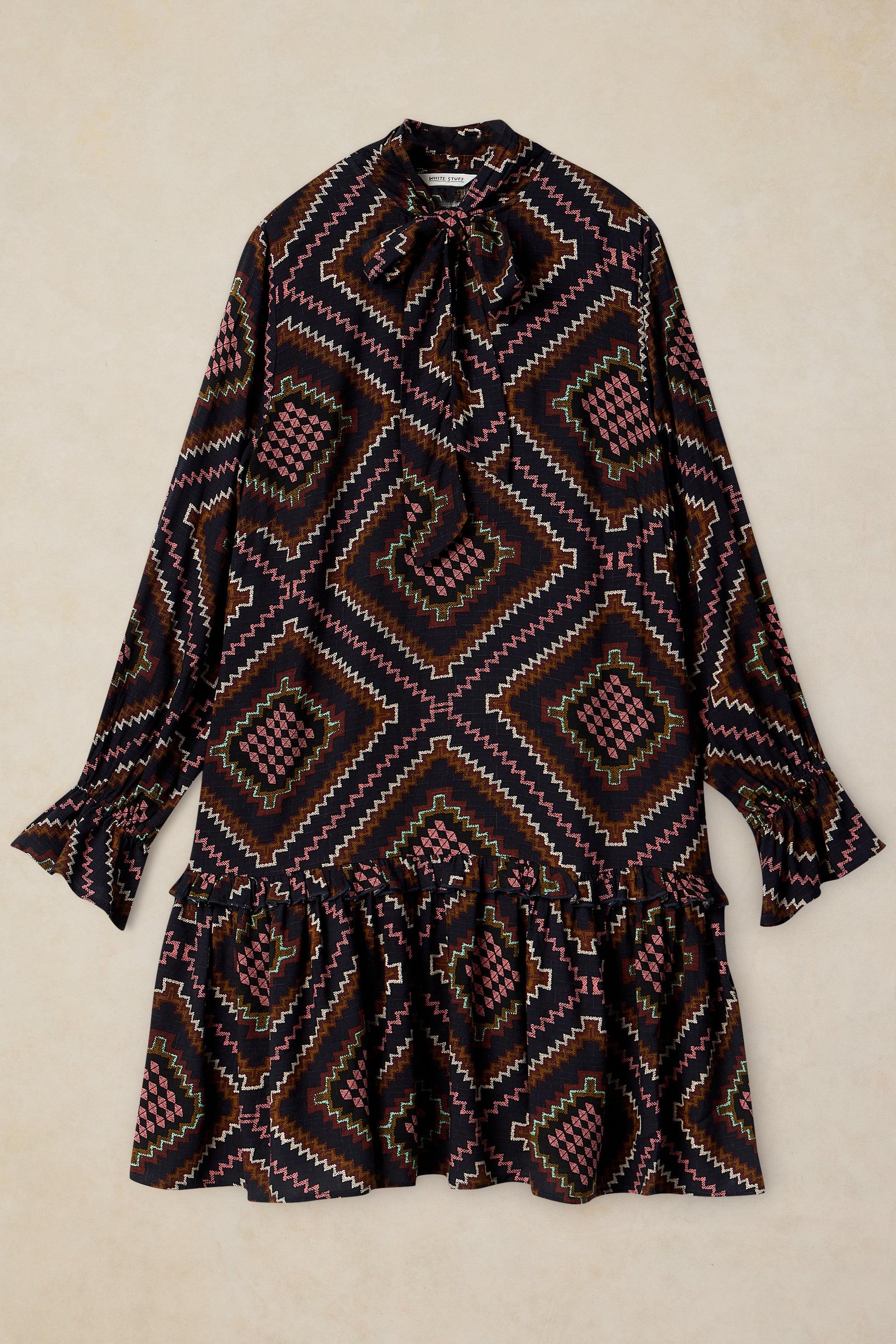 A long-sleeve mini dress with a geometric diamond pattern in black, brown, and pink, featuring a tie-neck detail and a ruffled hem, laid flat against a beige background.