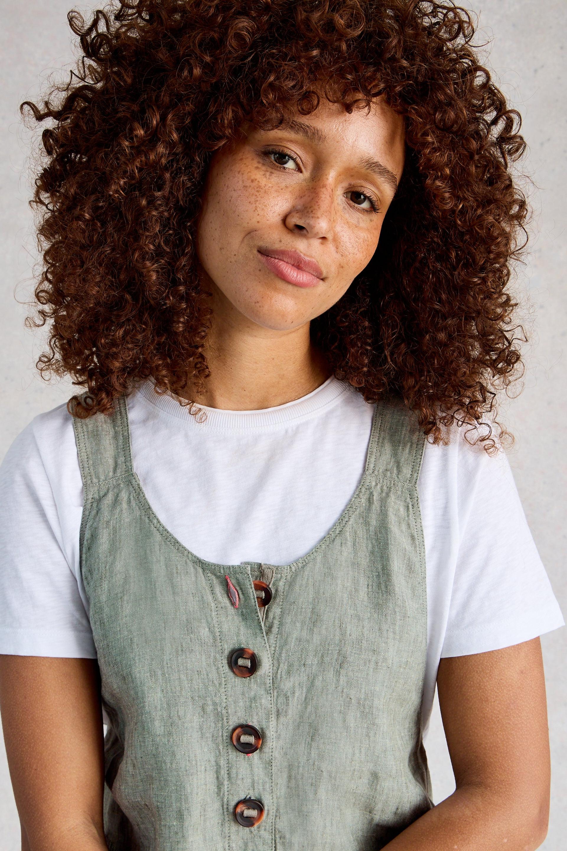 Woman with curly hair wearing a white shirt and green linen dungarees against a neutral background