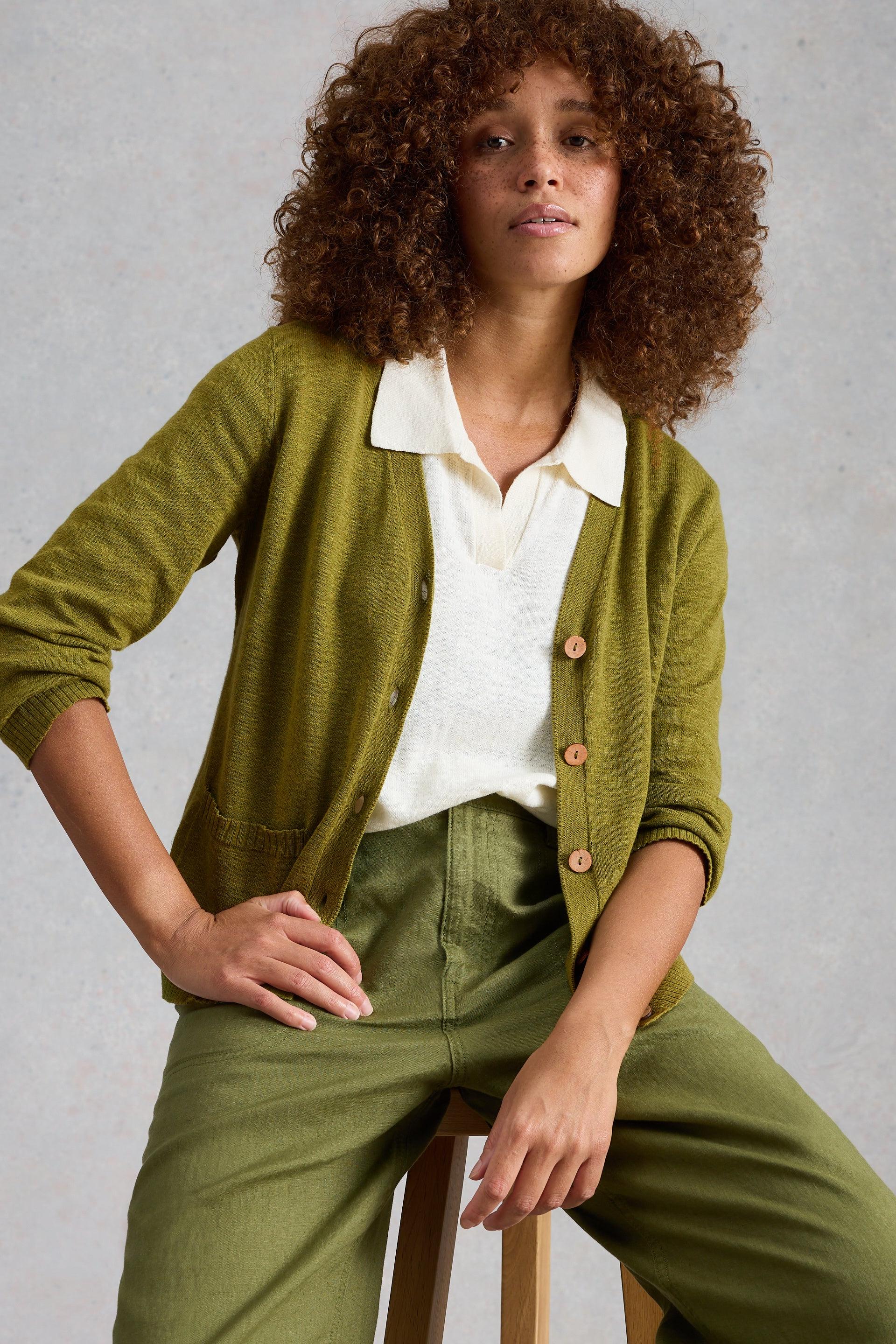 Woman wearing a green organic cotton cardigan over a white shirt and green pants, sitting on a stool against a gray background.