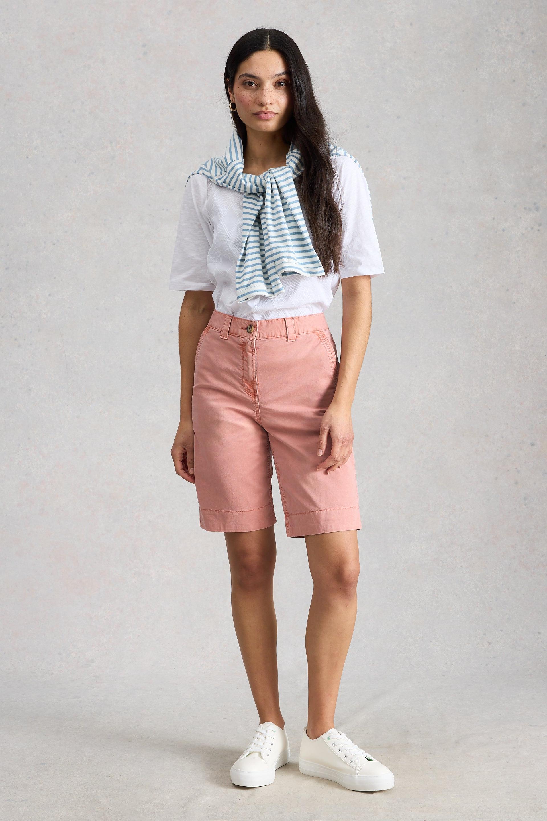 Woman wearing a white shirt, pink organic cotton shorts, and white sneakers on a plain background