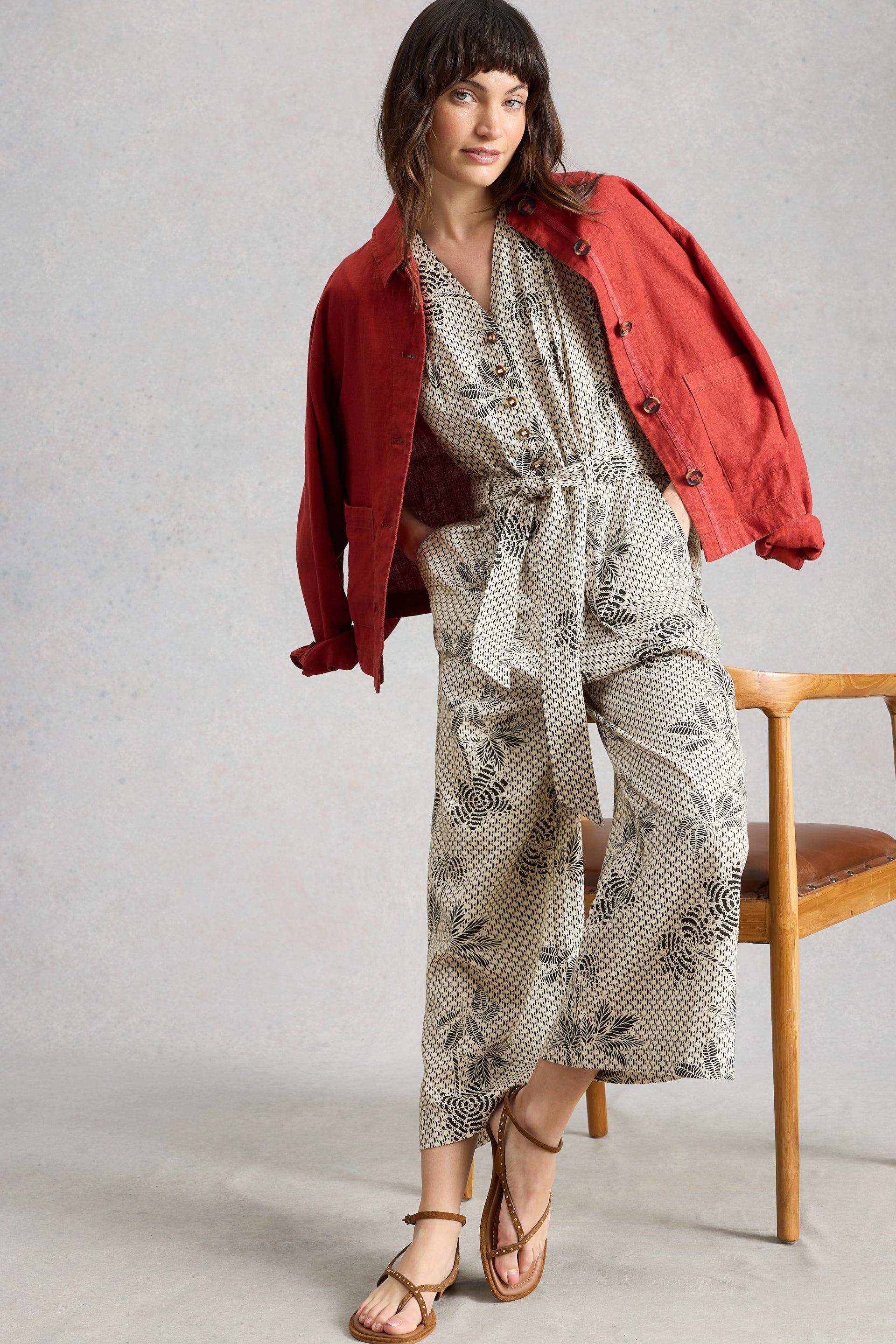 Woman wearing a red jacket over a patterned linen jumpsuit sitting on a wooden chair.