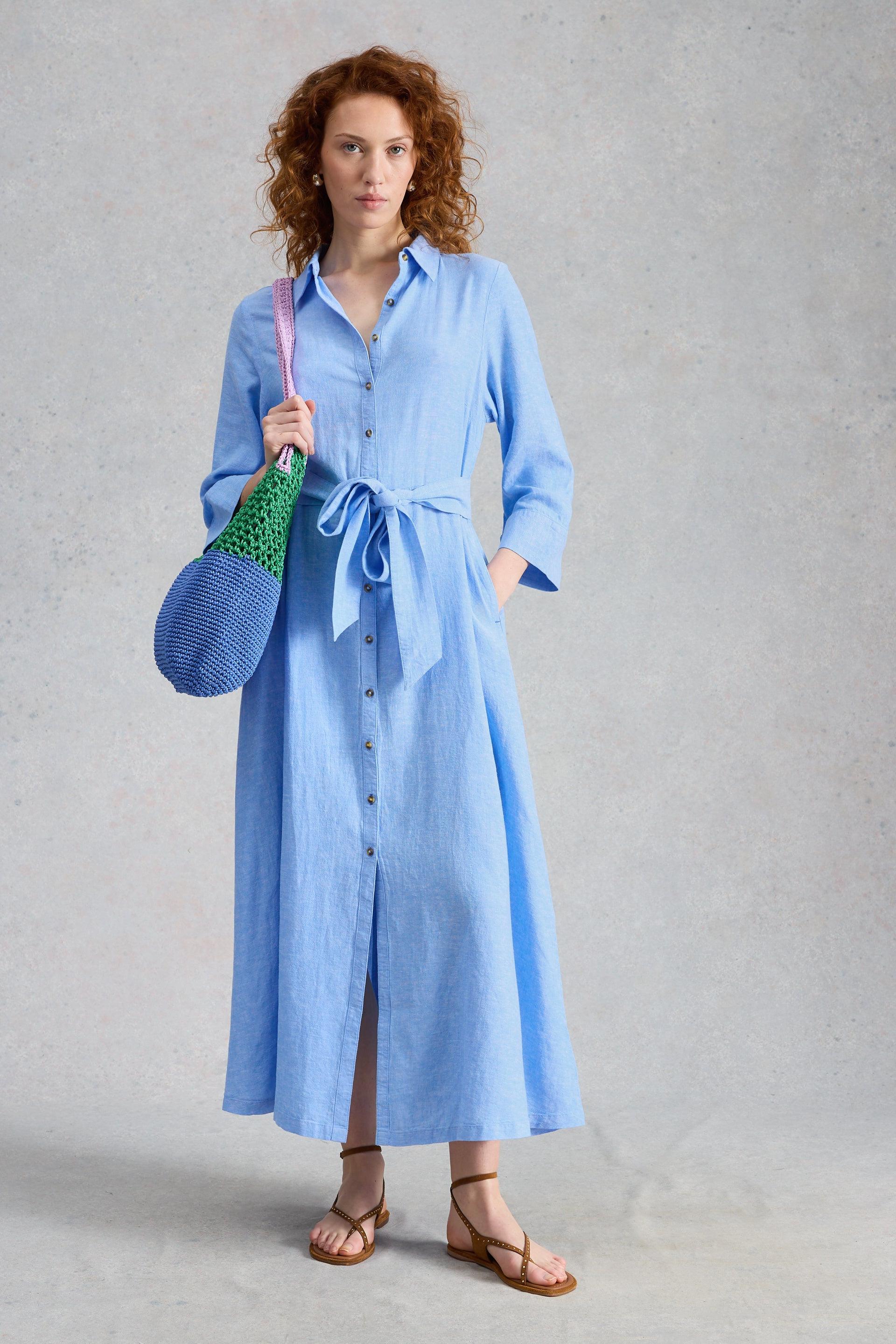 Woman wearing a light blue shirt dress holding a crochet bag against a gray background