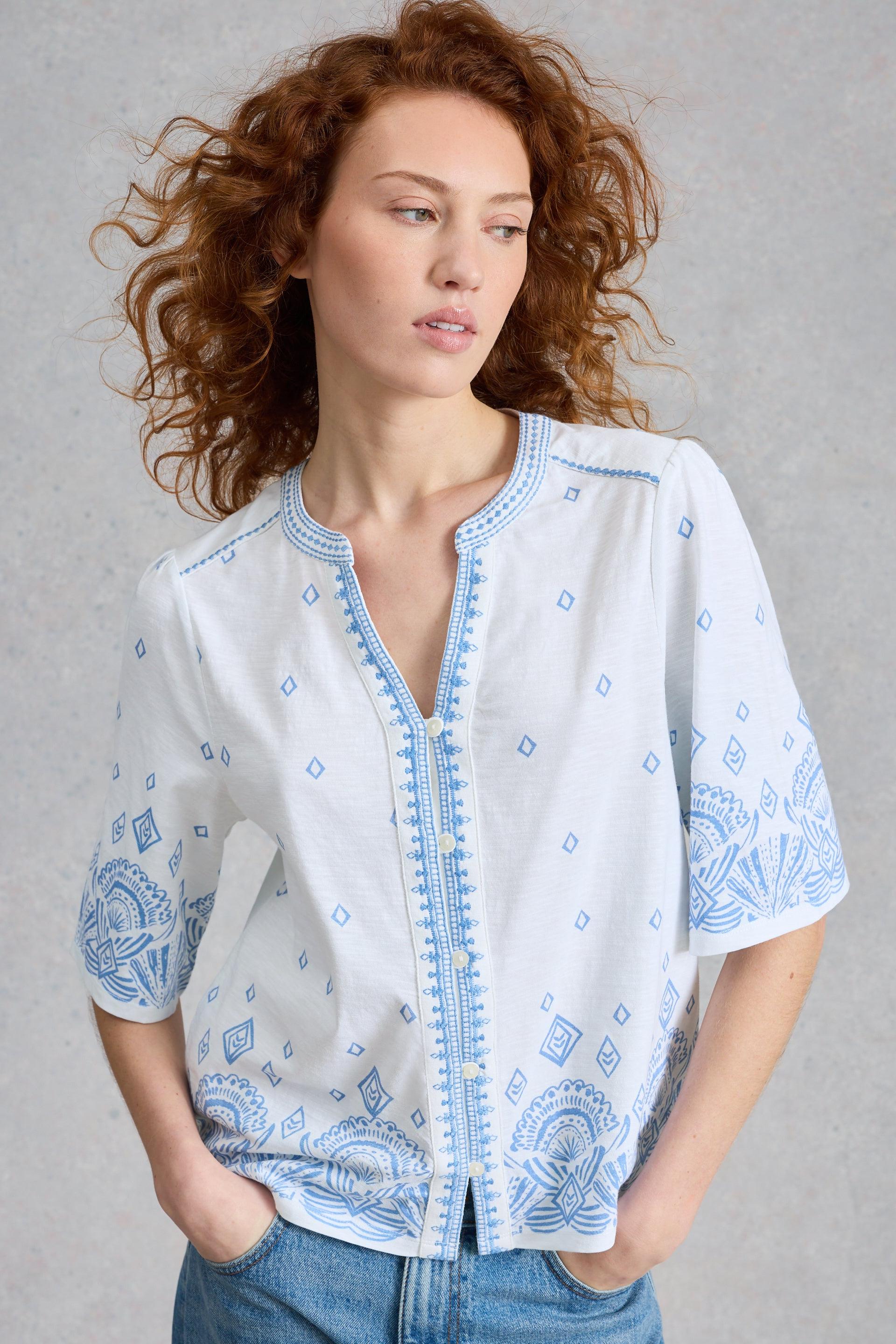 Woman wearing a white organic cotton blouse with blue patterns and blue jeans against a gray background