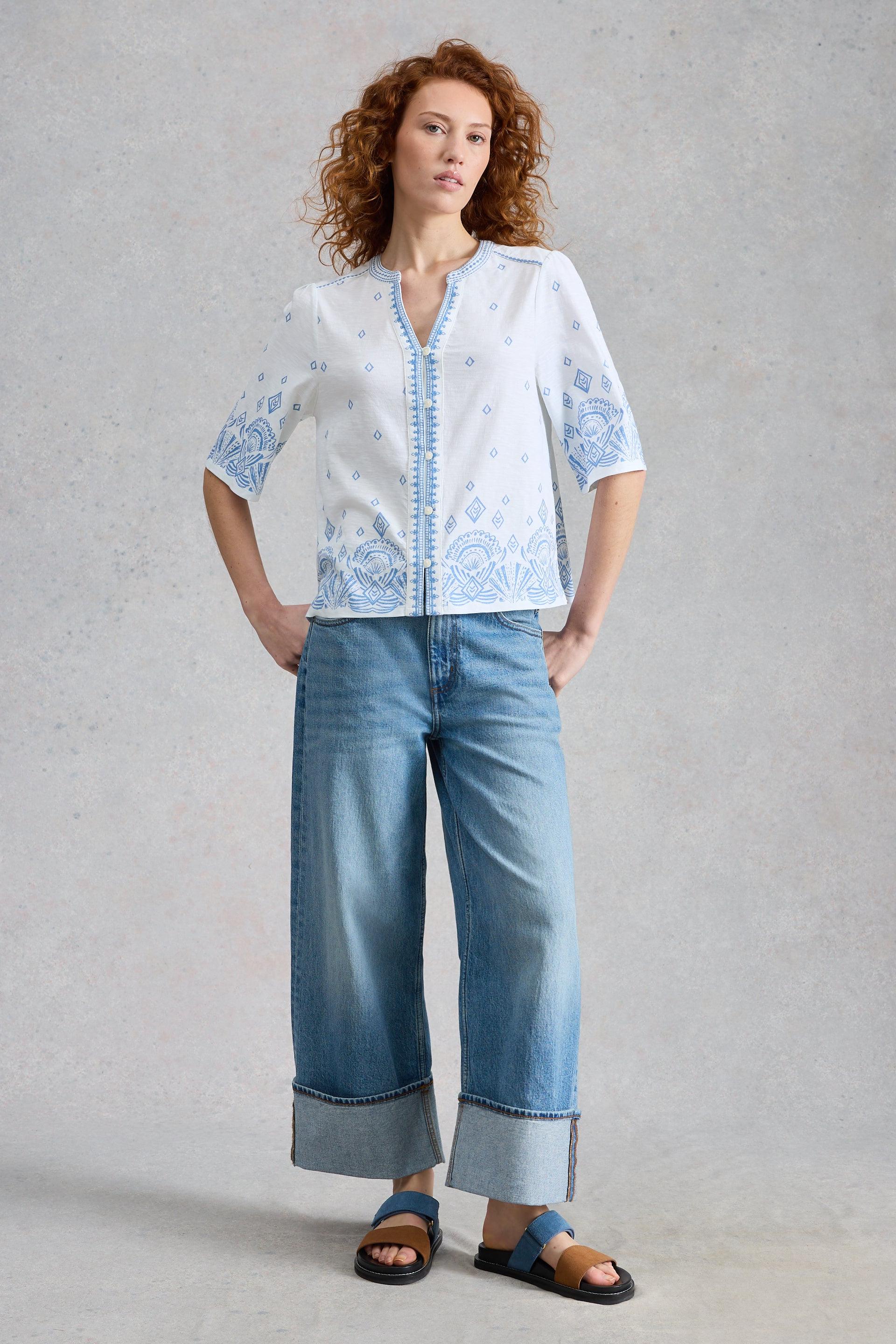 Woman wearing a white White Stuff blouse with blue patterns and wide-leg jeans on a plain background