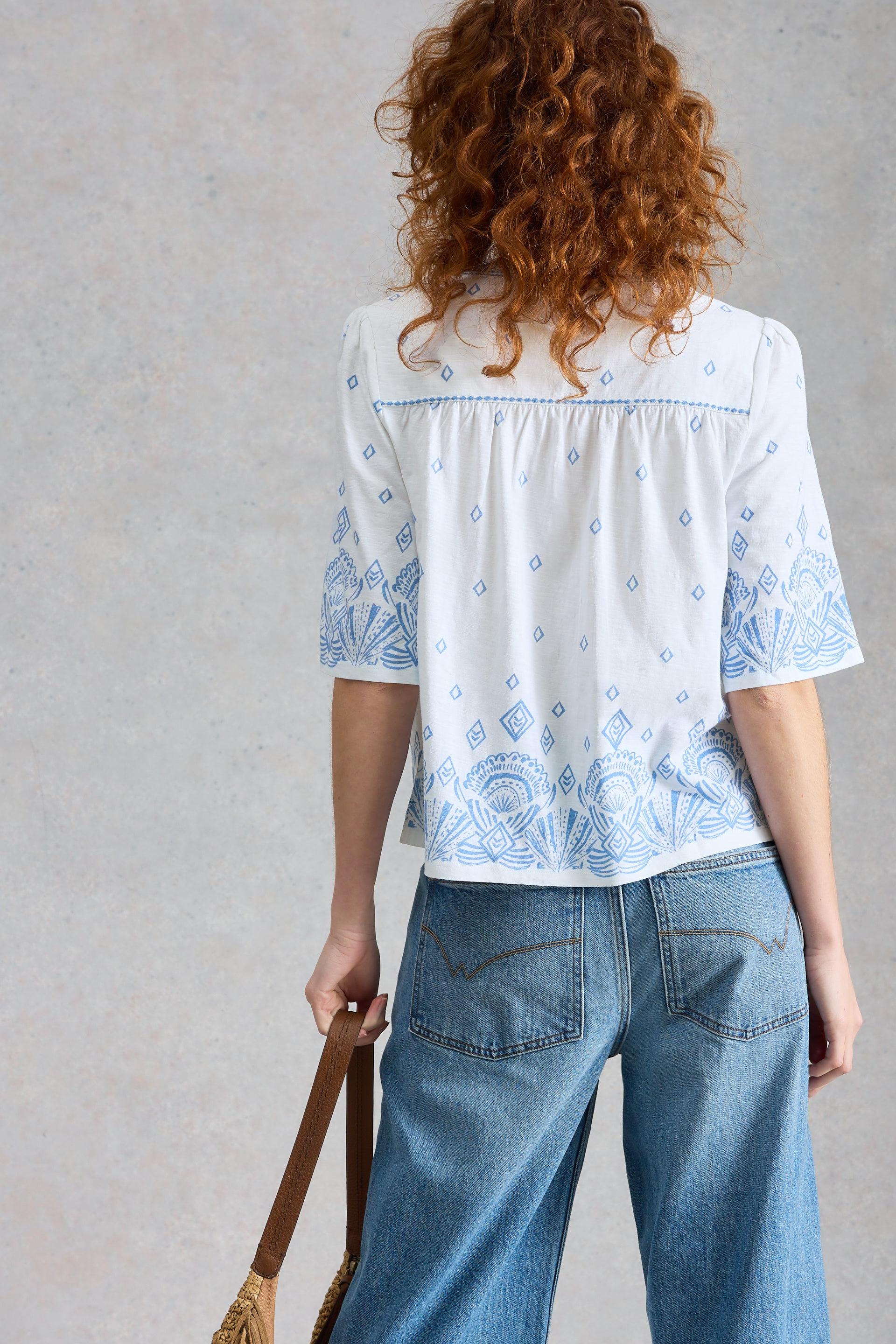 Person wearing a white blouse with blue patterns and blue jeans against a gray background