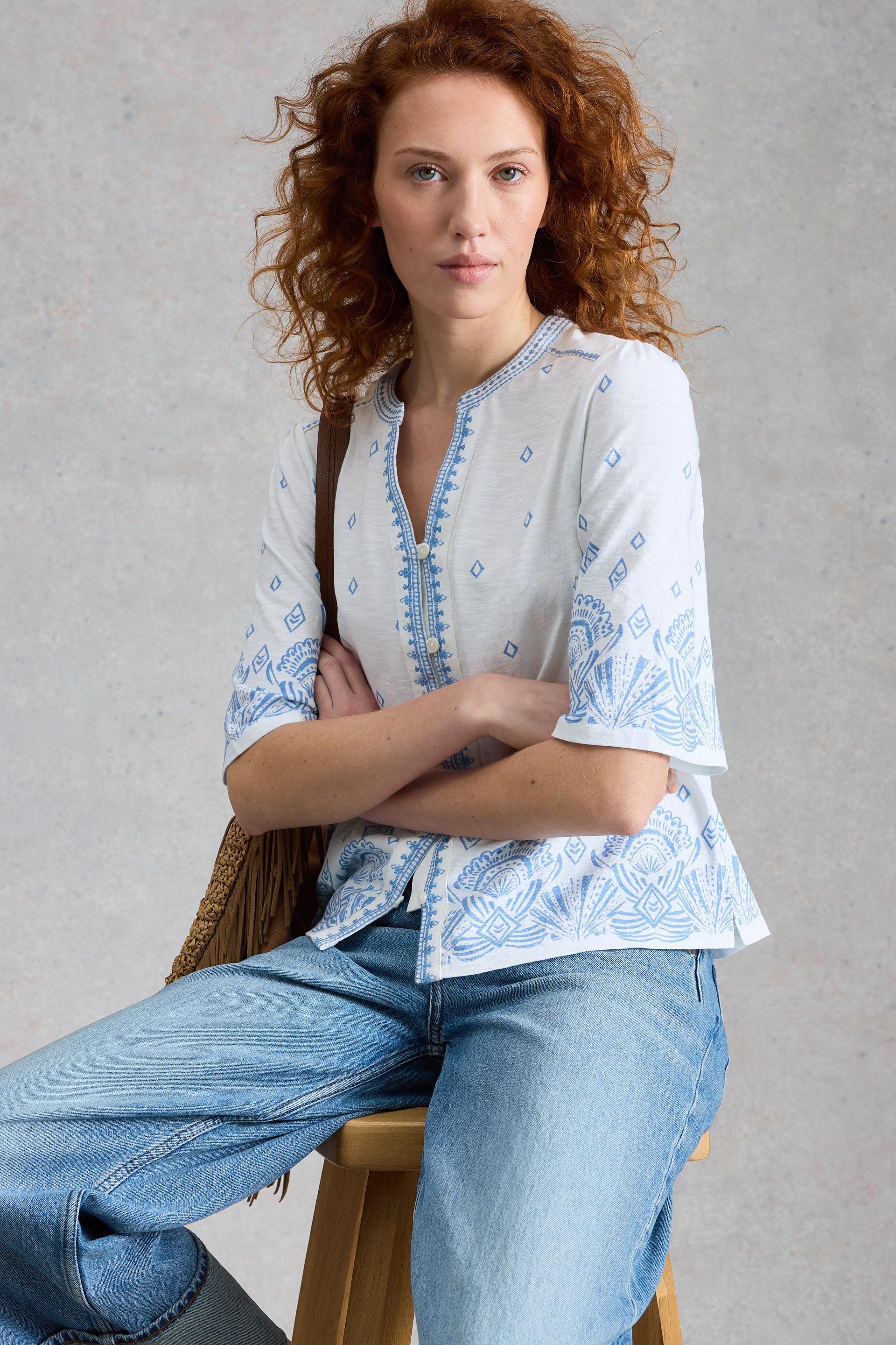 Woman wearing a white blouse with blue patterns and blue jeans, sitting on a wooden stool against a gray background.