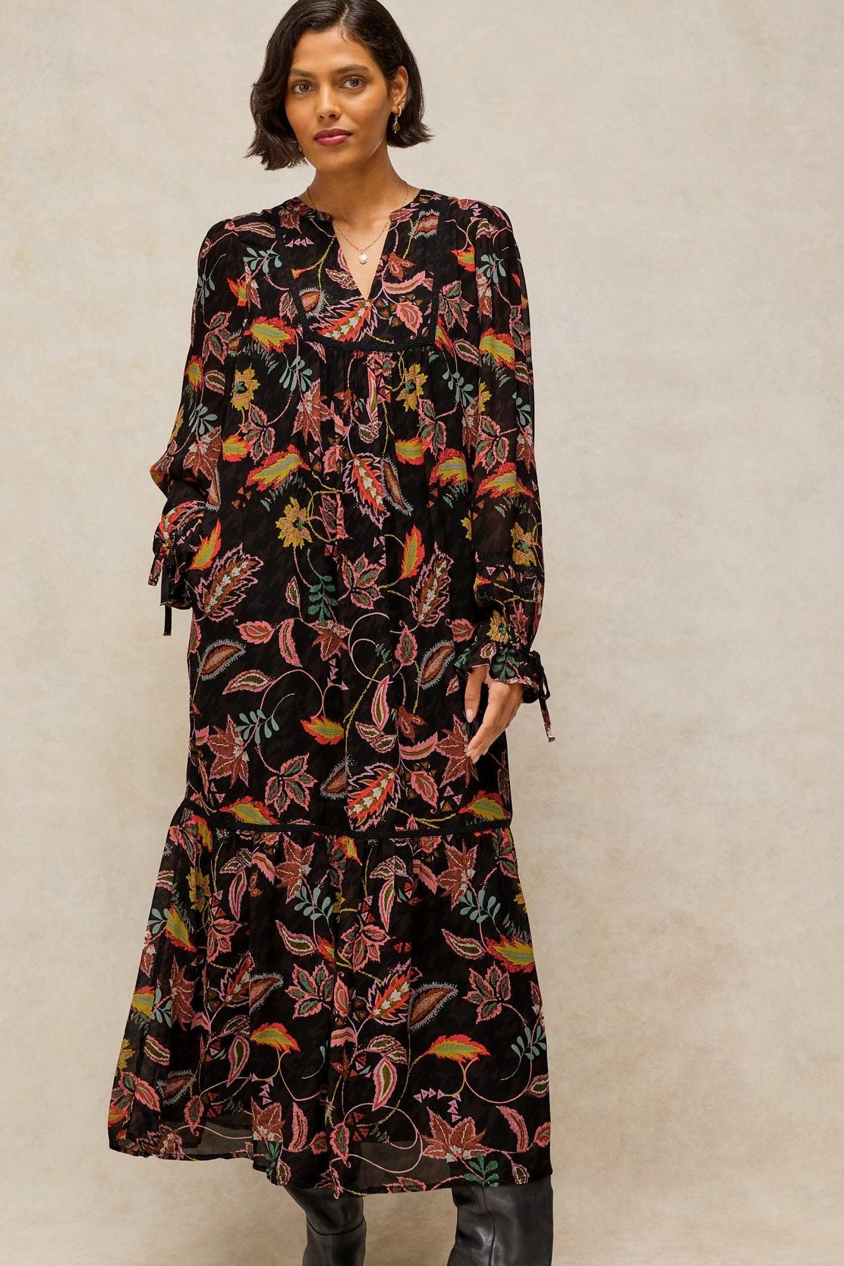 Woman wearing a long black floral White Stuff viscose evening dress with ruffled sleeves against a neutral background.