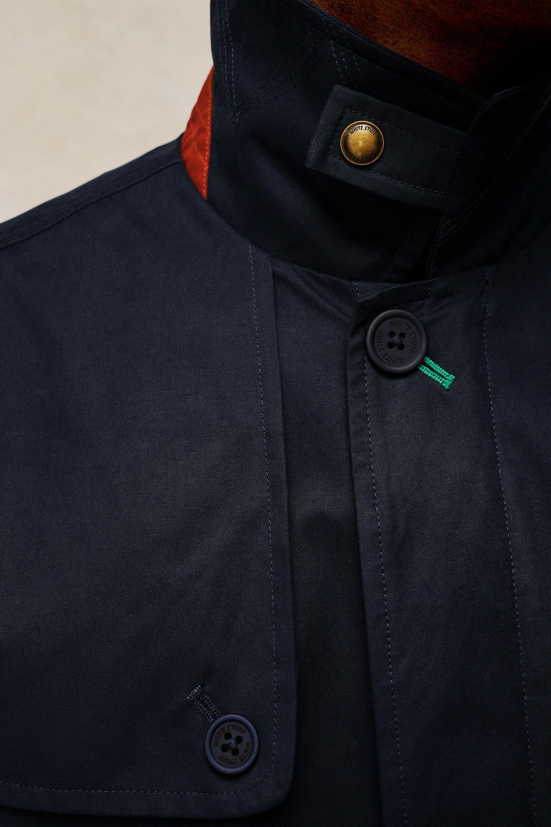 Close-up of a dark navy jacket with a high collar, a gold button, and a small green embroidered logo on the chest.