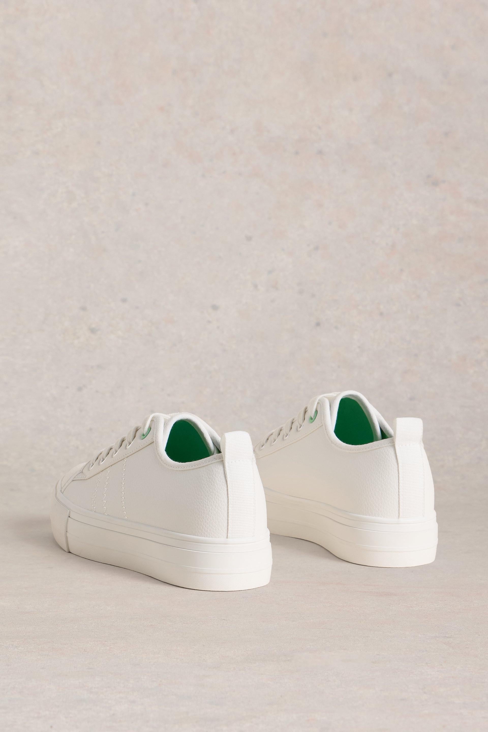 A pair of clean white minimalist sneakers with green lining displayed against a textured neutral background.