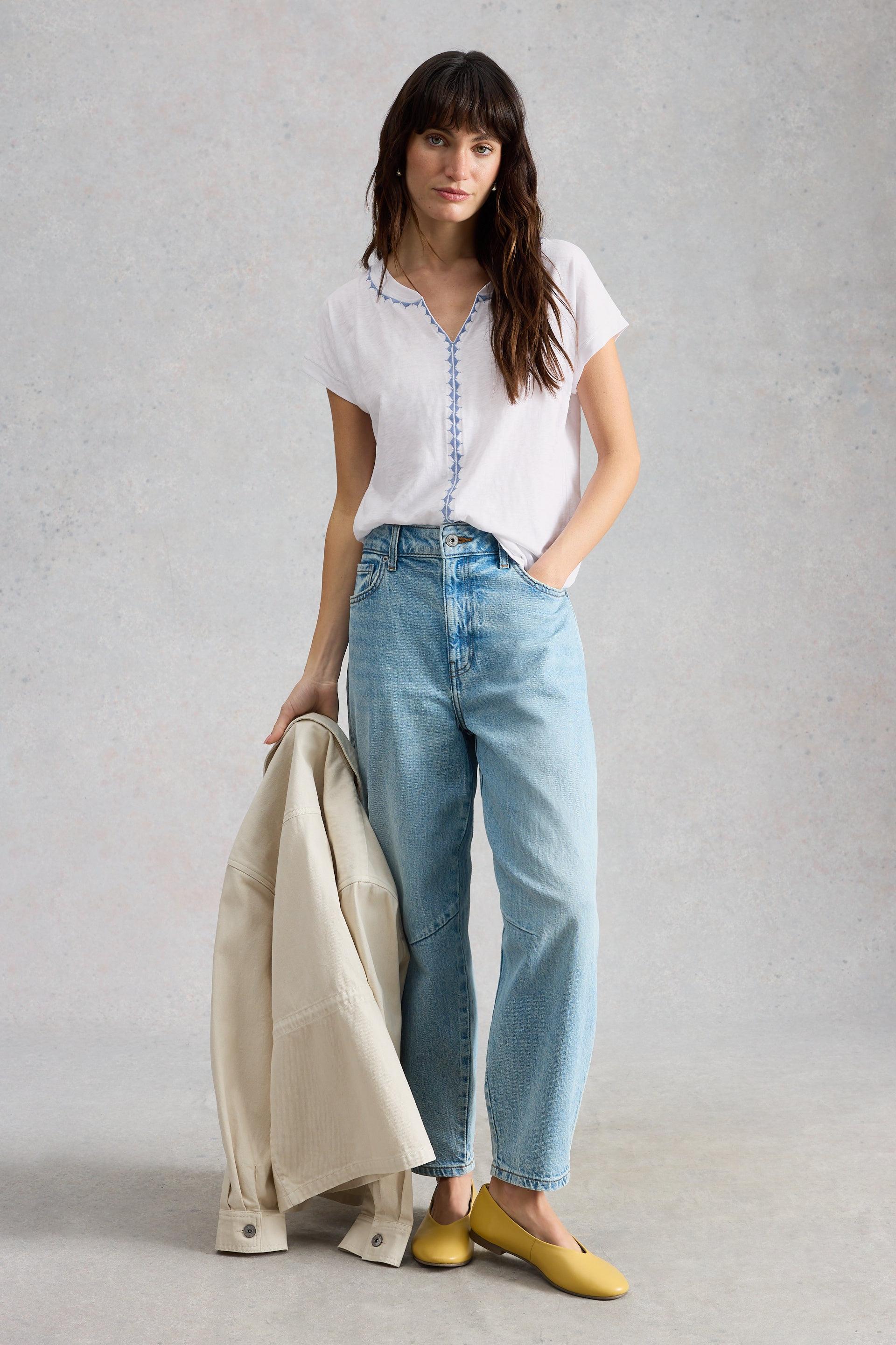 Woman wearing a white cotton shirt, blue jeans, and yellow shoes holding a beige jacket against a gray background