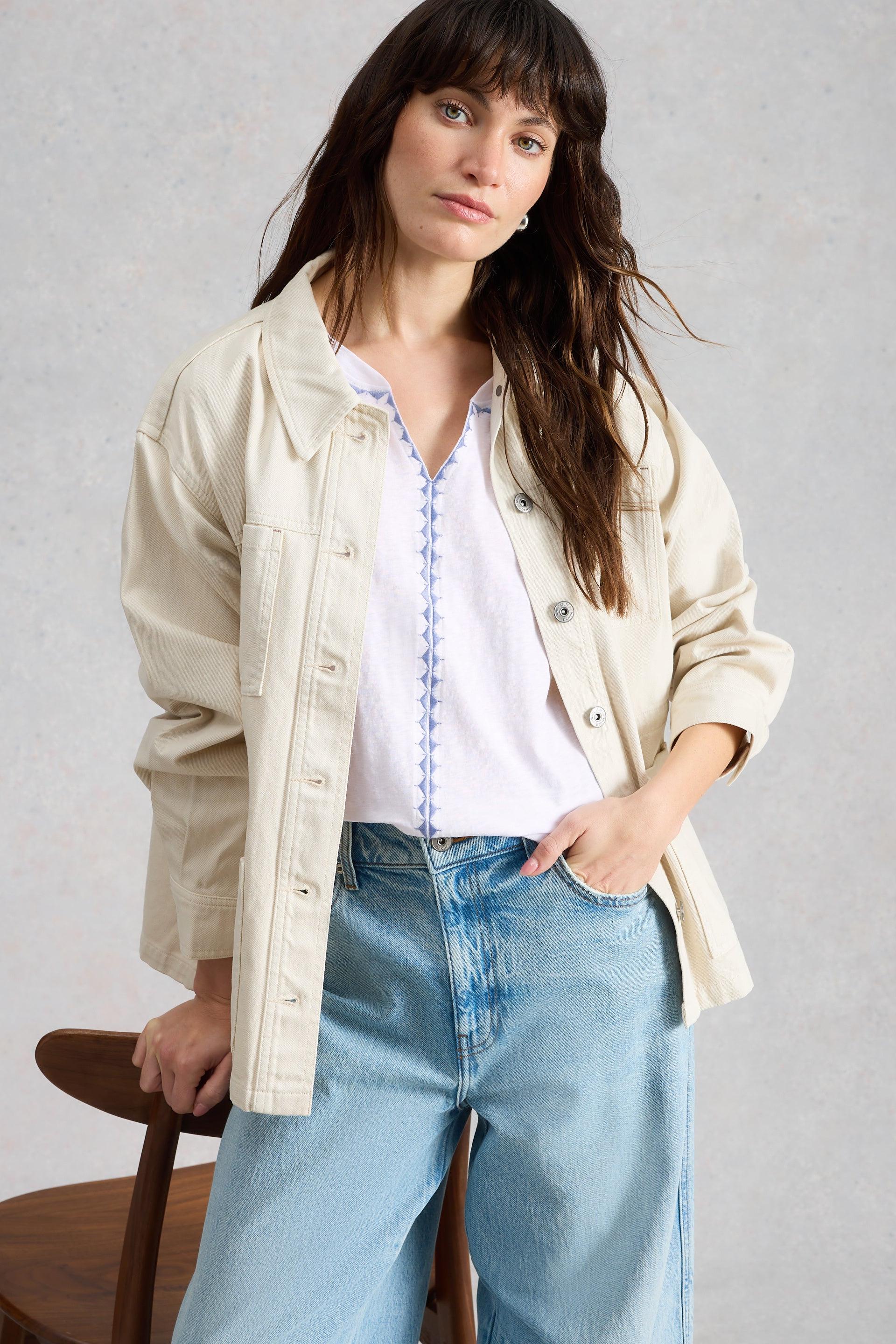 Woman wearing a beige jacket, white t-shirt, and blue jeans against a gray background