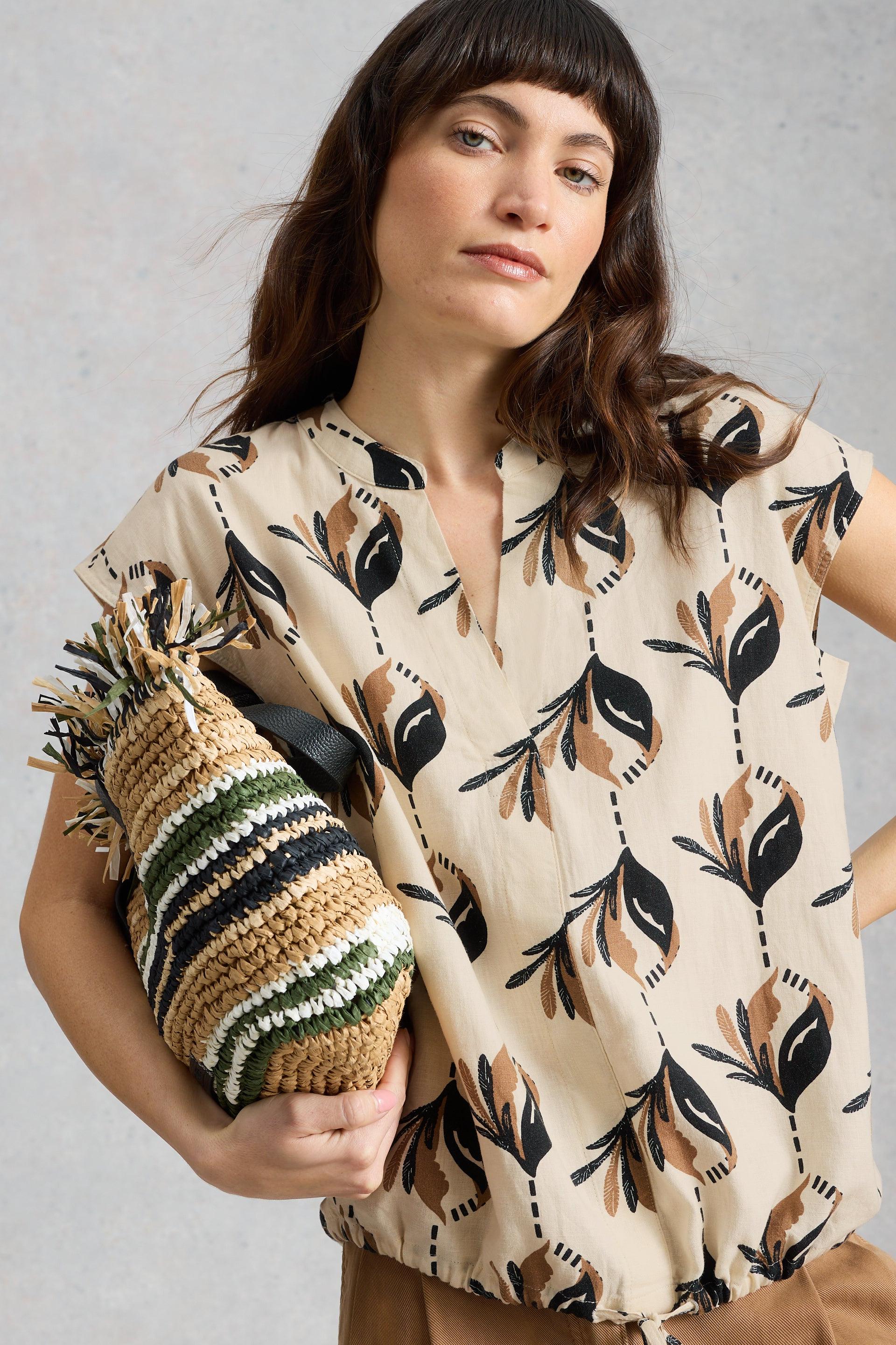 Woman wearing a beige linen blouse with leaf pattern holding a woven bag against a gray background
