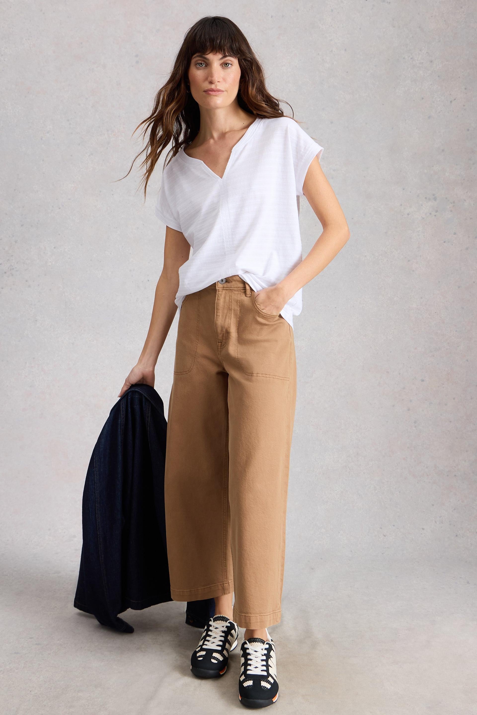 Woman wearing a white short sleeve top and tan pants holding a black jacket against a plain background