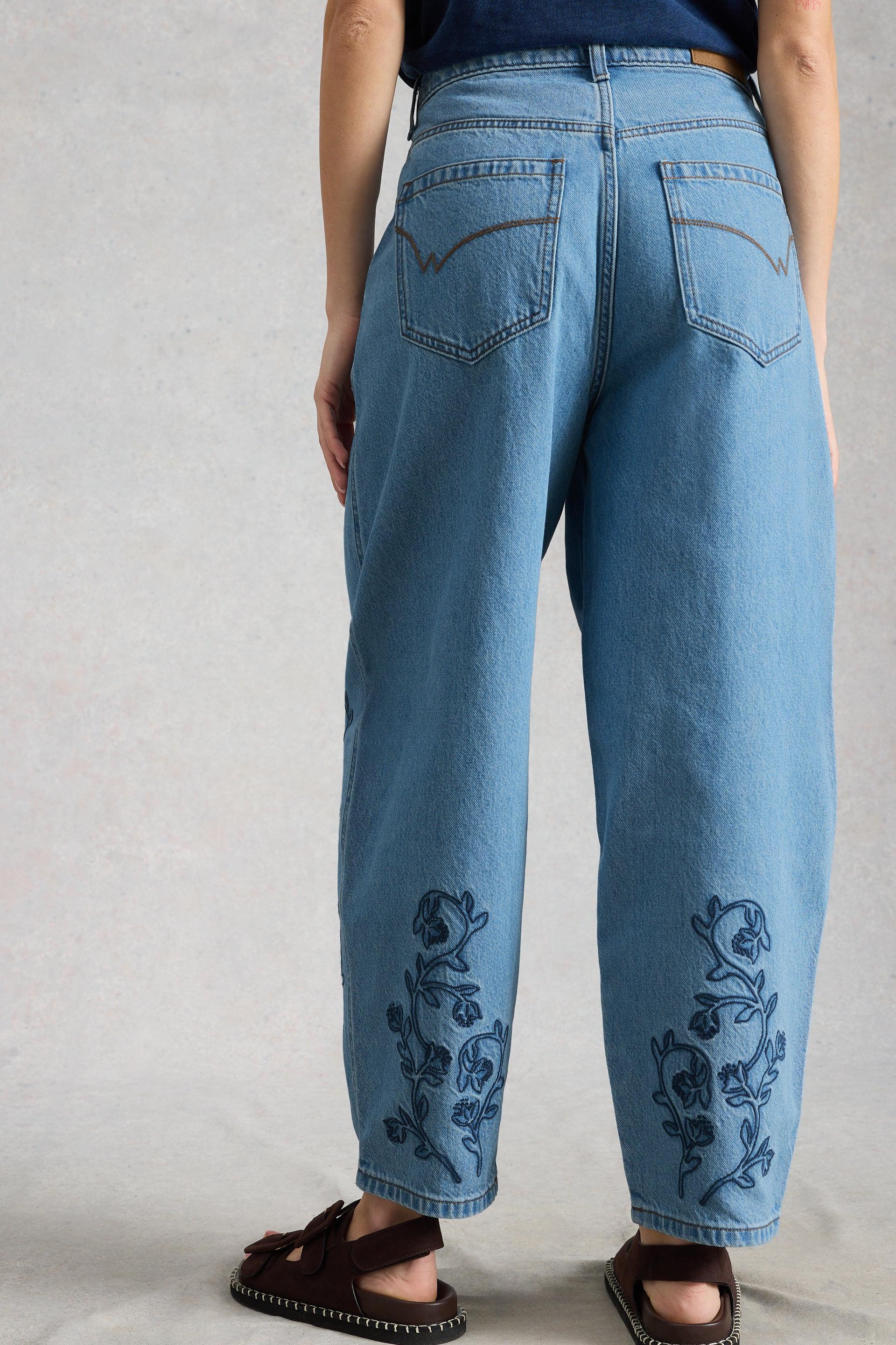 Model wearing blue barrel leg jeans with floral embroidery on a plain background