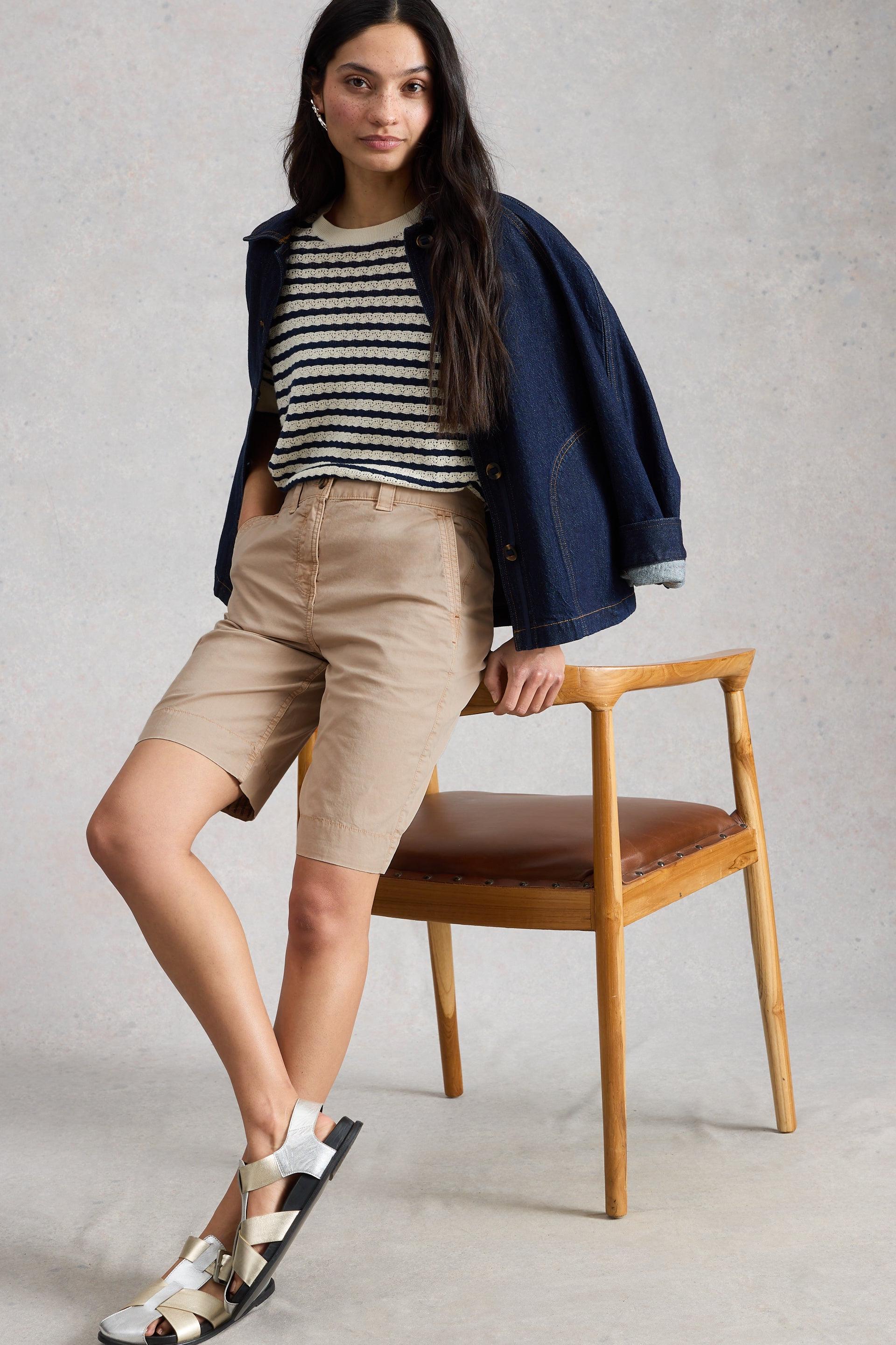 Woman sitting on a wooden chair wearing beige White Stuff shorts, a striped shirt, and a navy jacket.