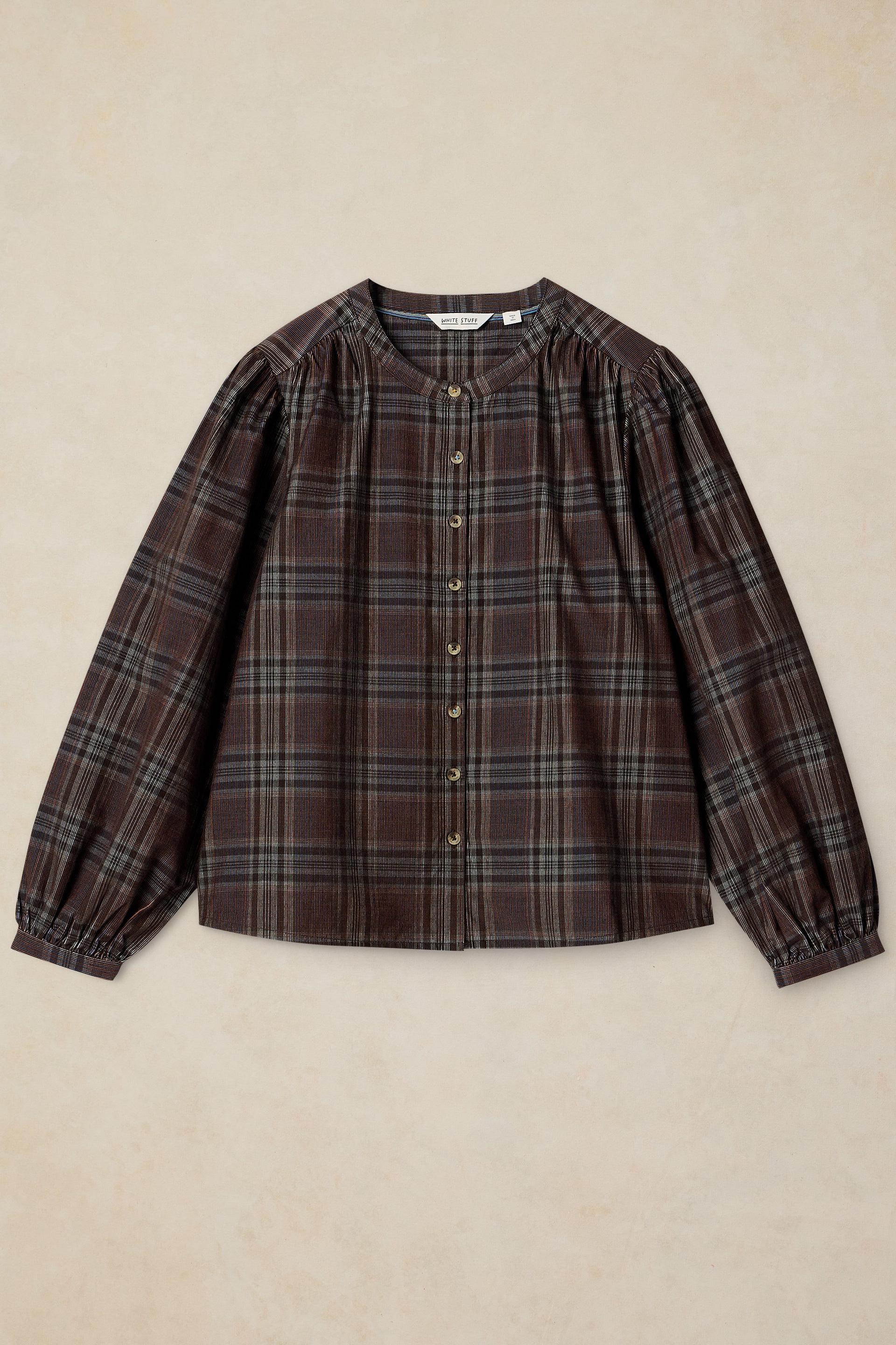 A long-sleeve button-up shirt in a dark brown and black plaid pattern, featuring a gathered round neckline and elasticized cuffs, laid flat on a textured beige background.