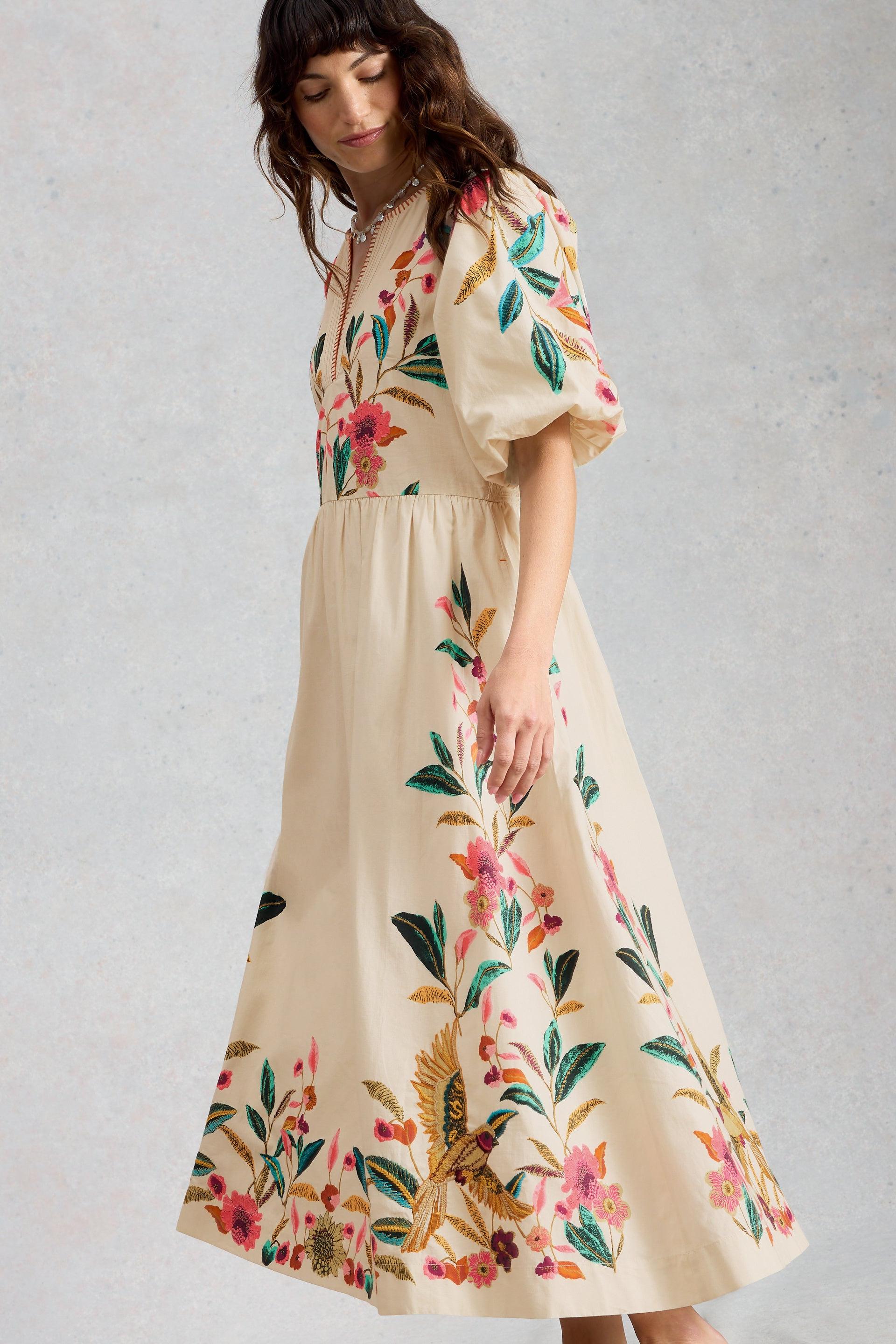 A woman wearing a cream-colored organic cotton midi occasion dress with vibrant floral embroidery.