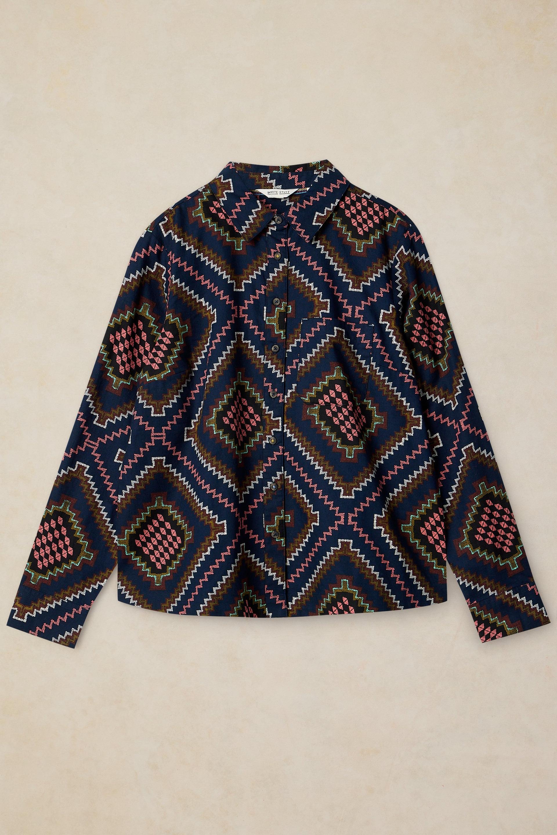 A long-sleeve button-up shirt with a dark navy blue base and a colorful geometric diamond pattern in pink, green, and beige, laid flat on a light beige background.