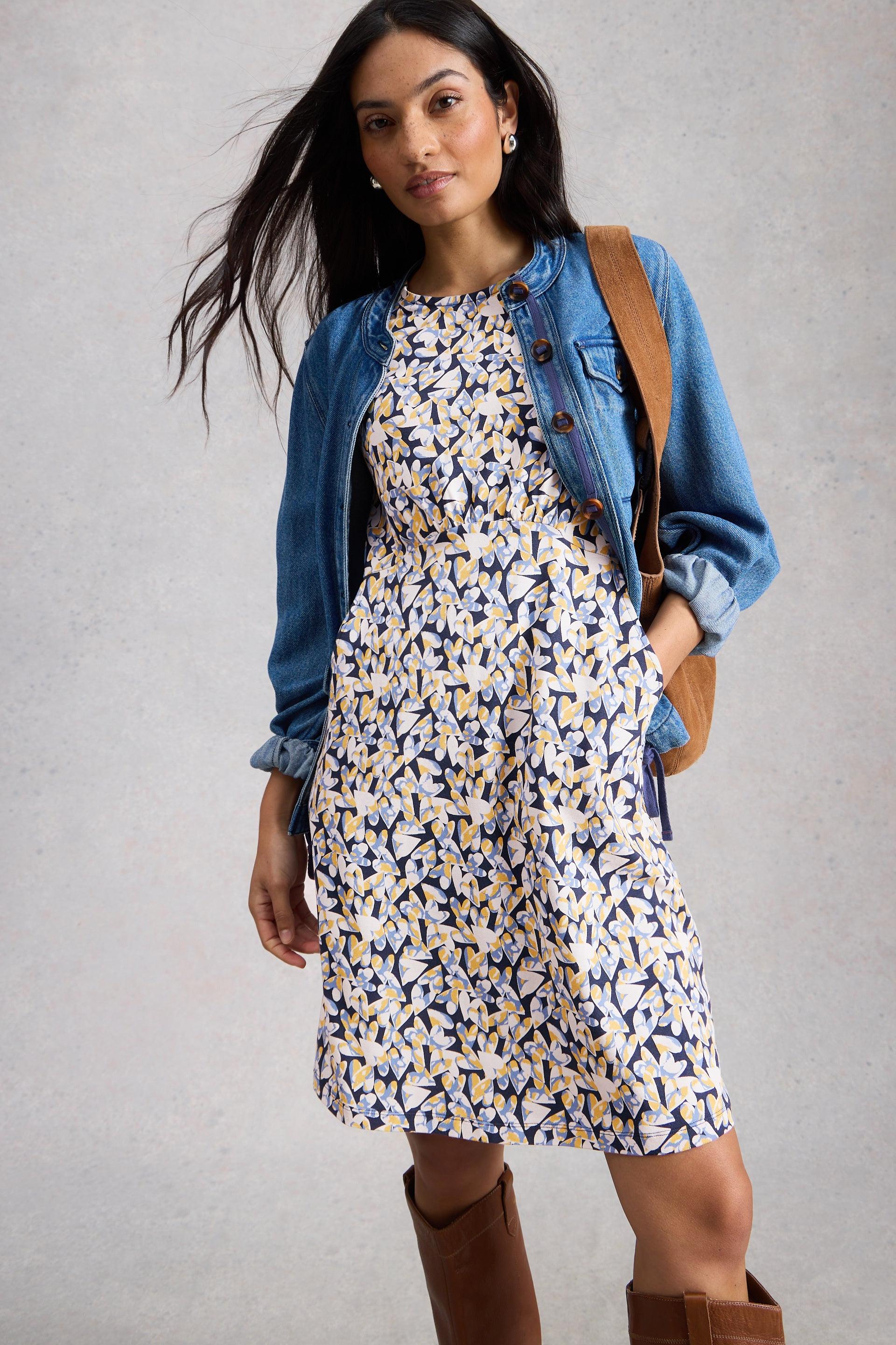 Woman wearing a floral organic cotton dress with a denim jacket and brown boots on a plain background