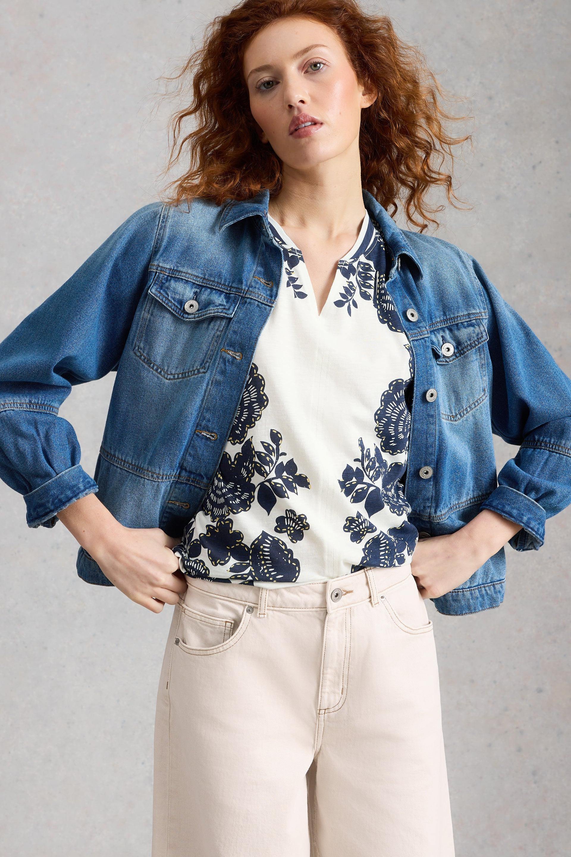 Woman wearing a denim jacket over a floral organic cotton blouse with beige pants on a gray background