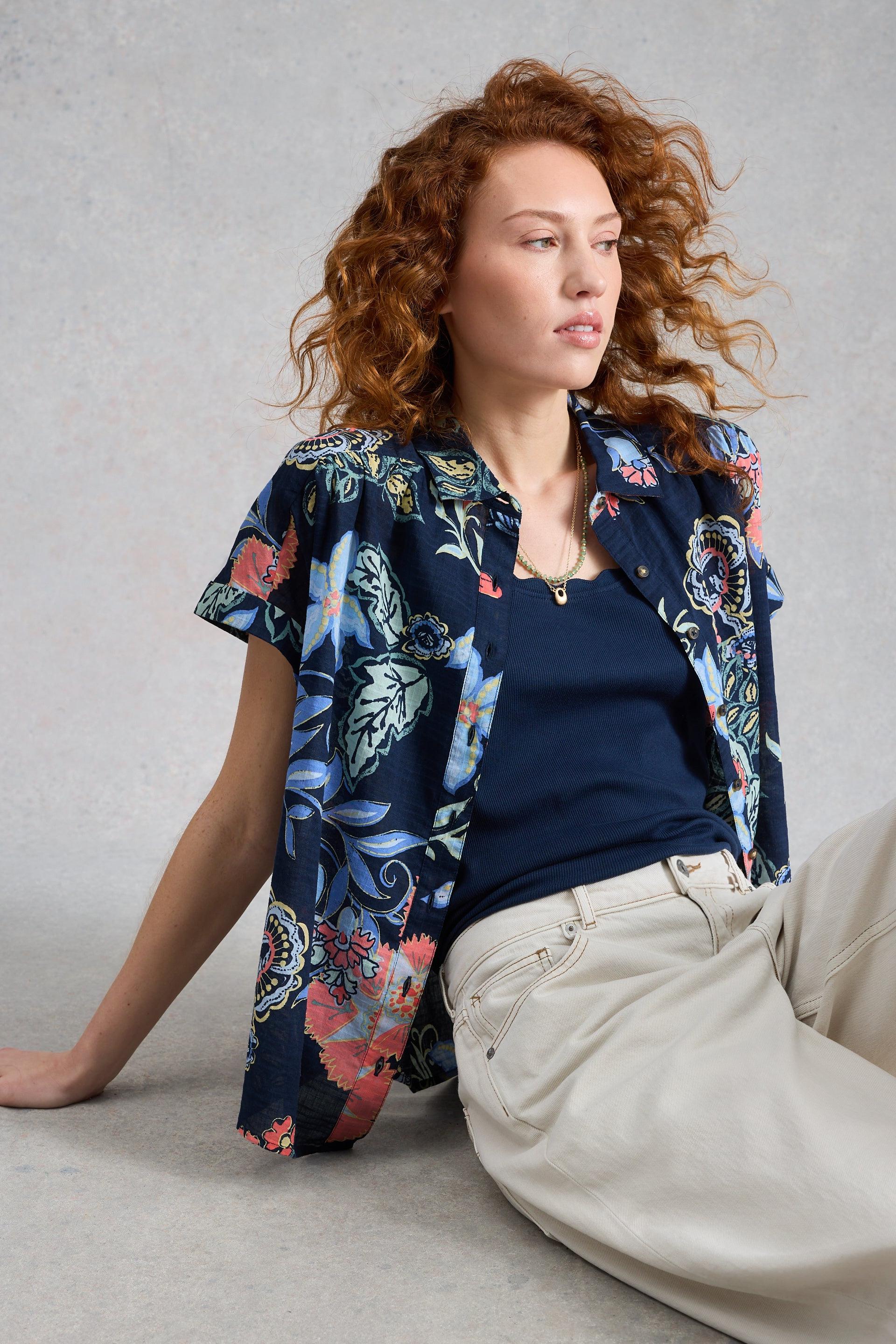 Woman wearing a floral patterned shirt over a dark top and light pants on a plain background