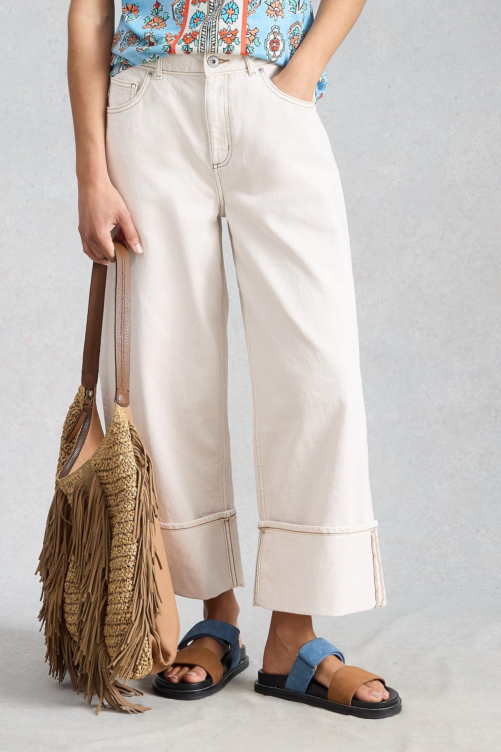 A model wearing white organic cotton trousers from White Stuff, accessorized with a woven fringe bag and flat sandals.