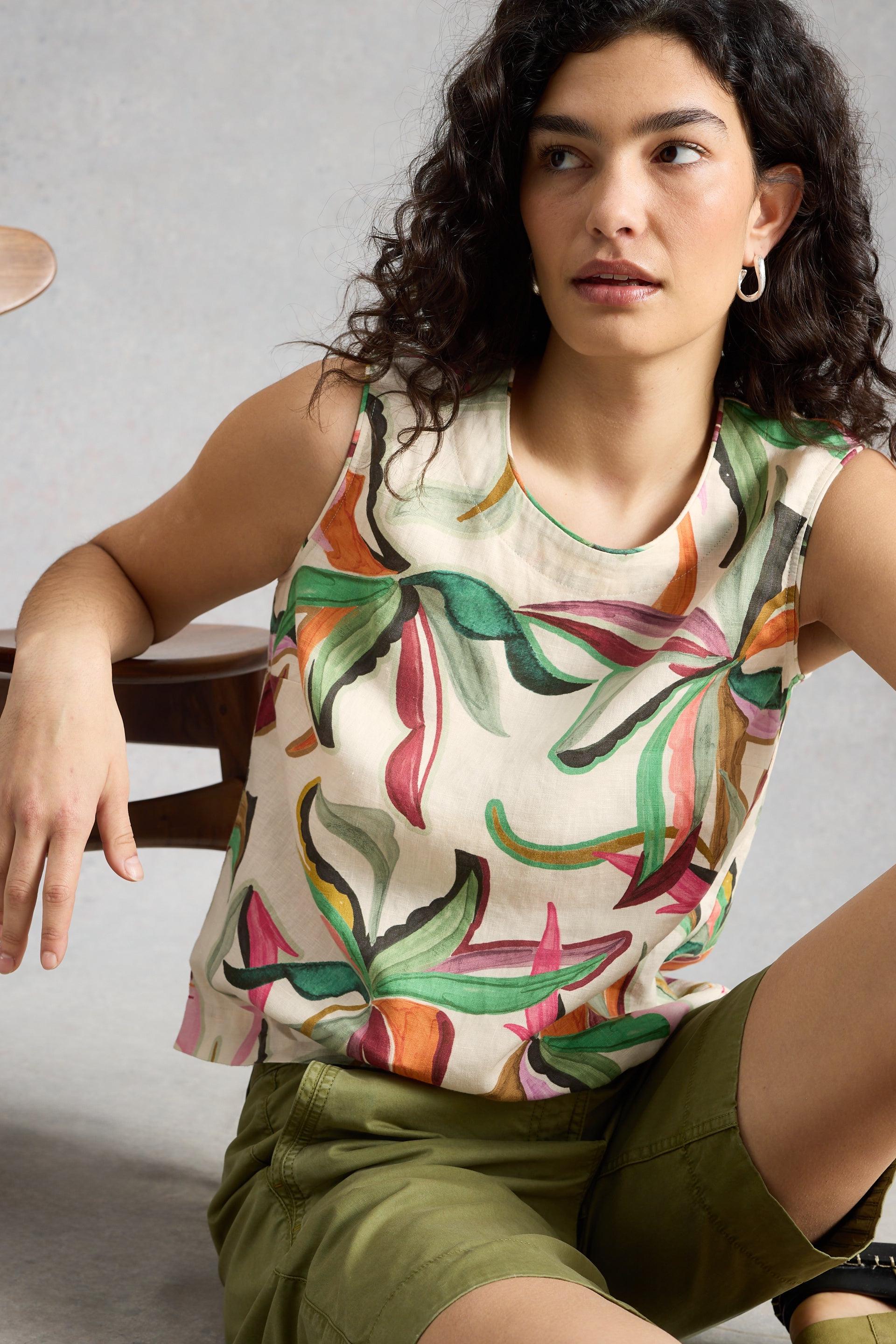 Woman wearing a sleeveless top with a colorful leaf pattern and green shorts, leaning on a chair.