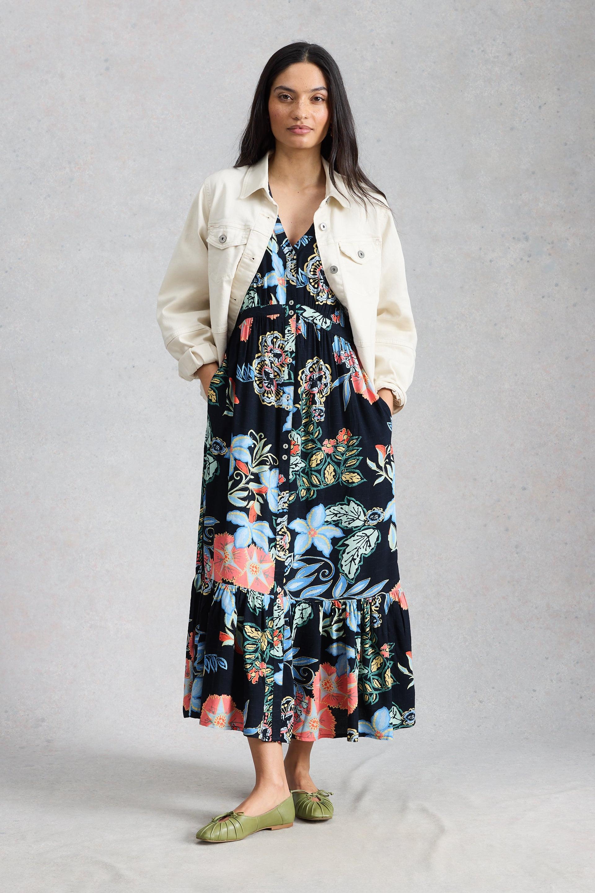 Woman wearing a floral White Stuff dress and cream denim jacket on a gray background