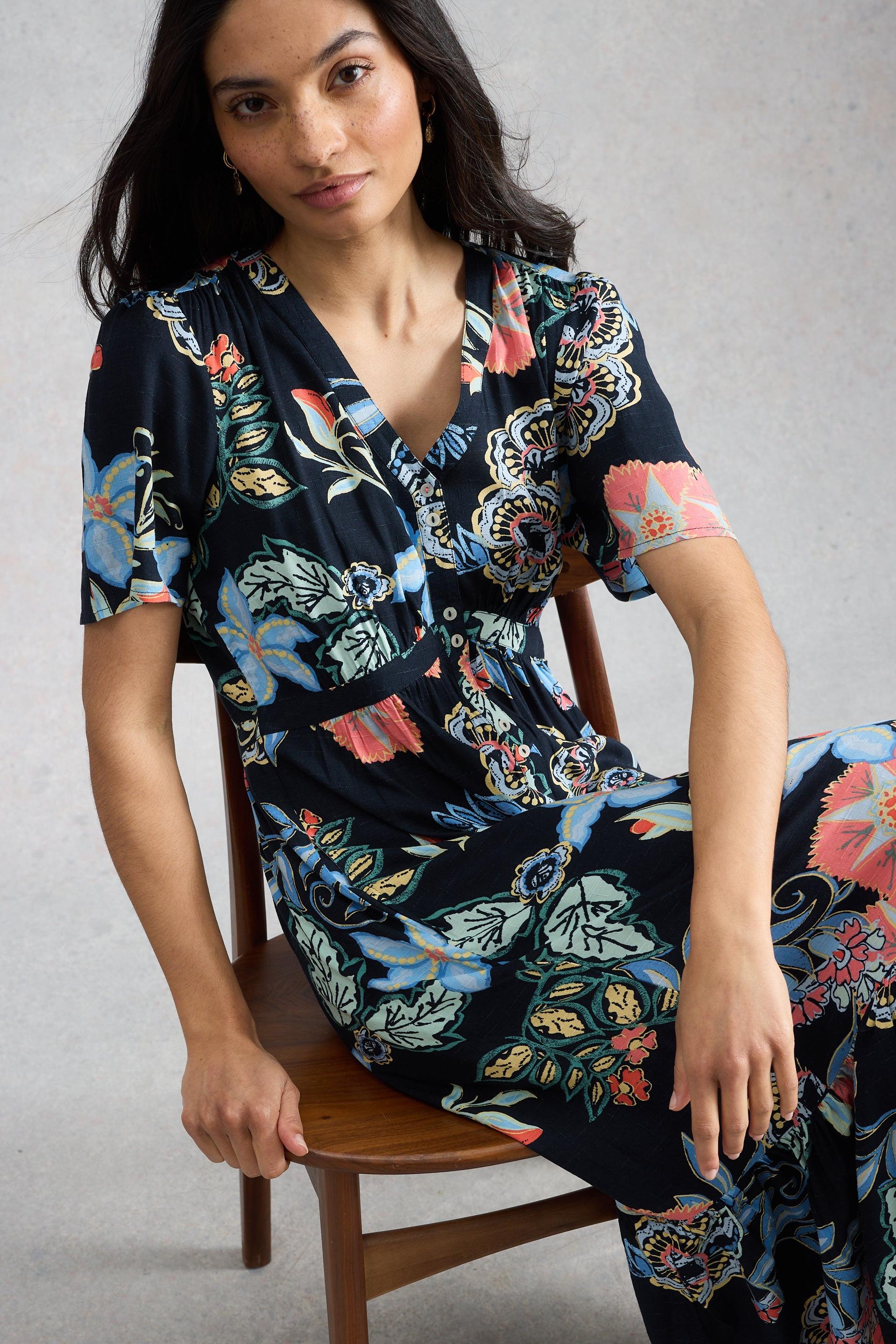 Woman wearing a floral White Stuff dress sitting on a chair against a plain background