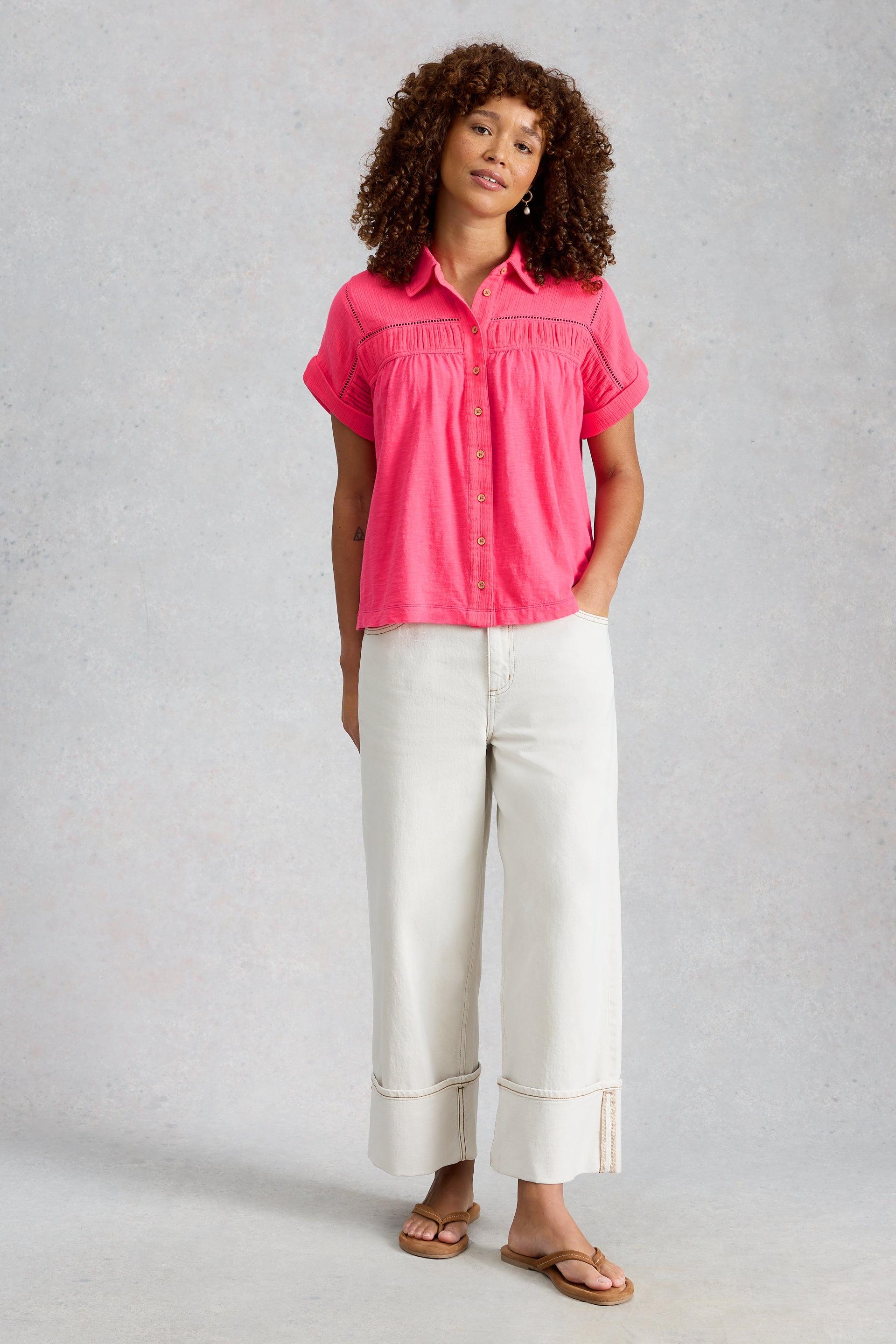 A woman wearing a vibrant pink shirt and off-white organic cotton trousers standing against a textured grey background.