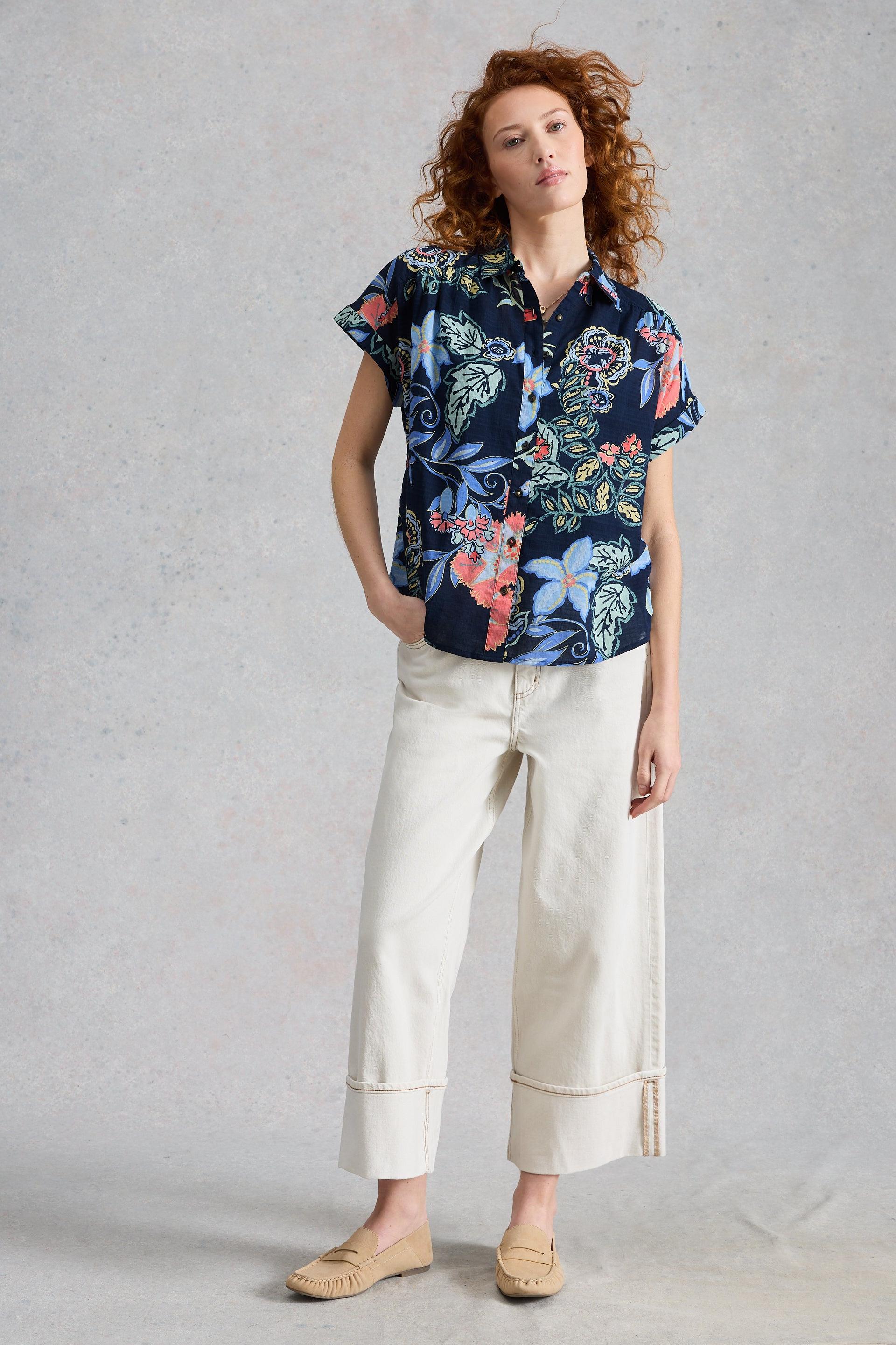 Woman wearing a floral White Stuff blouse and white pants against a gray background