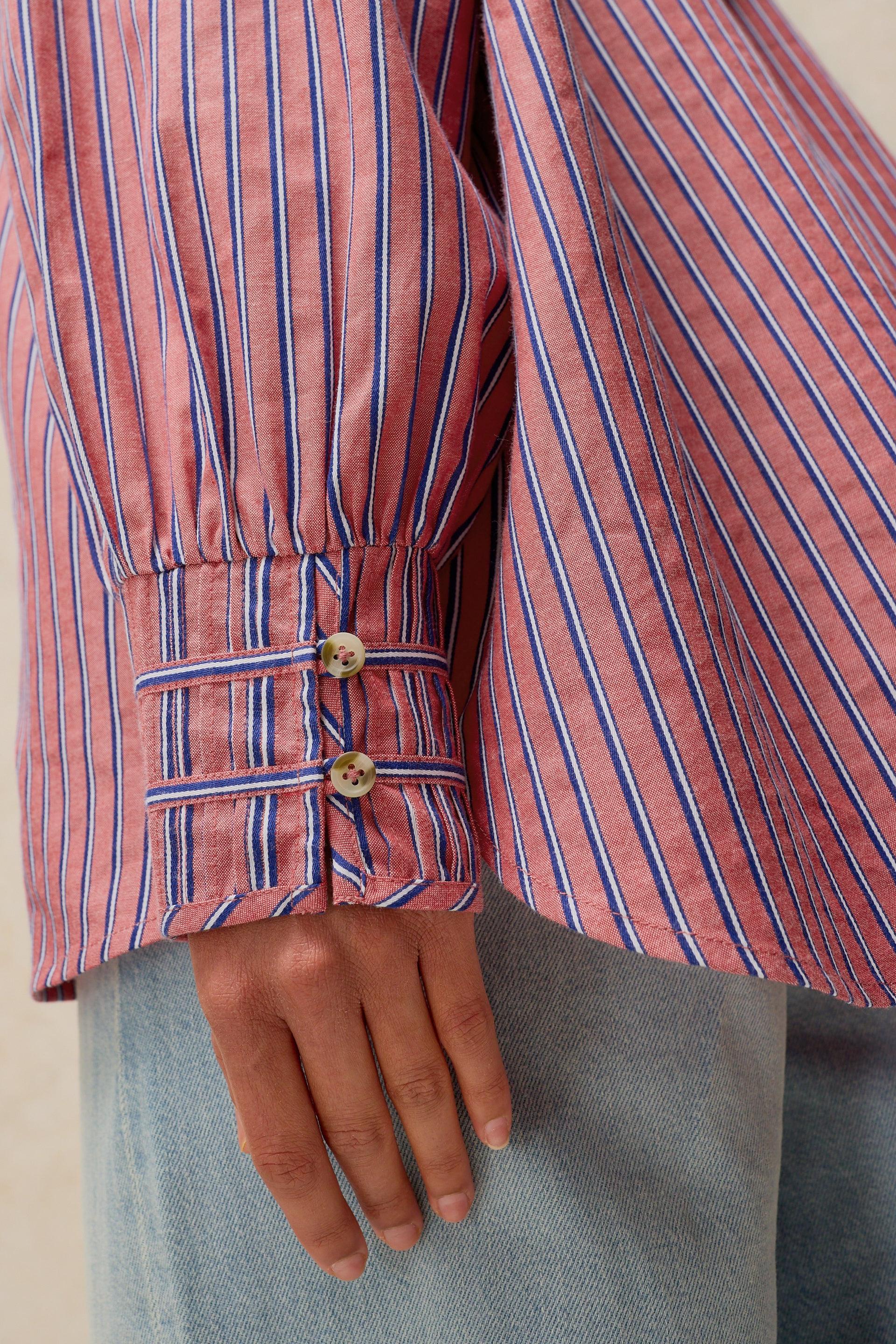 Close-up of a pink striped long-sleeve blouse with blue and white vertical pinstripes, featuring a gathered cuff with two decorative buttons, worn with light blue jeans.
