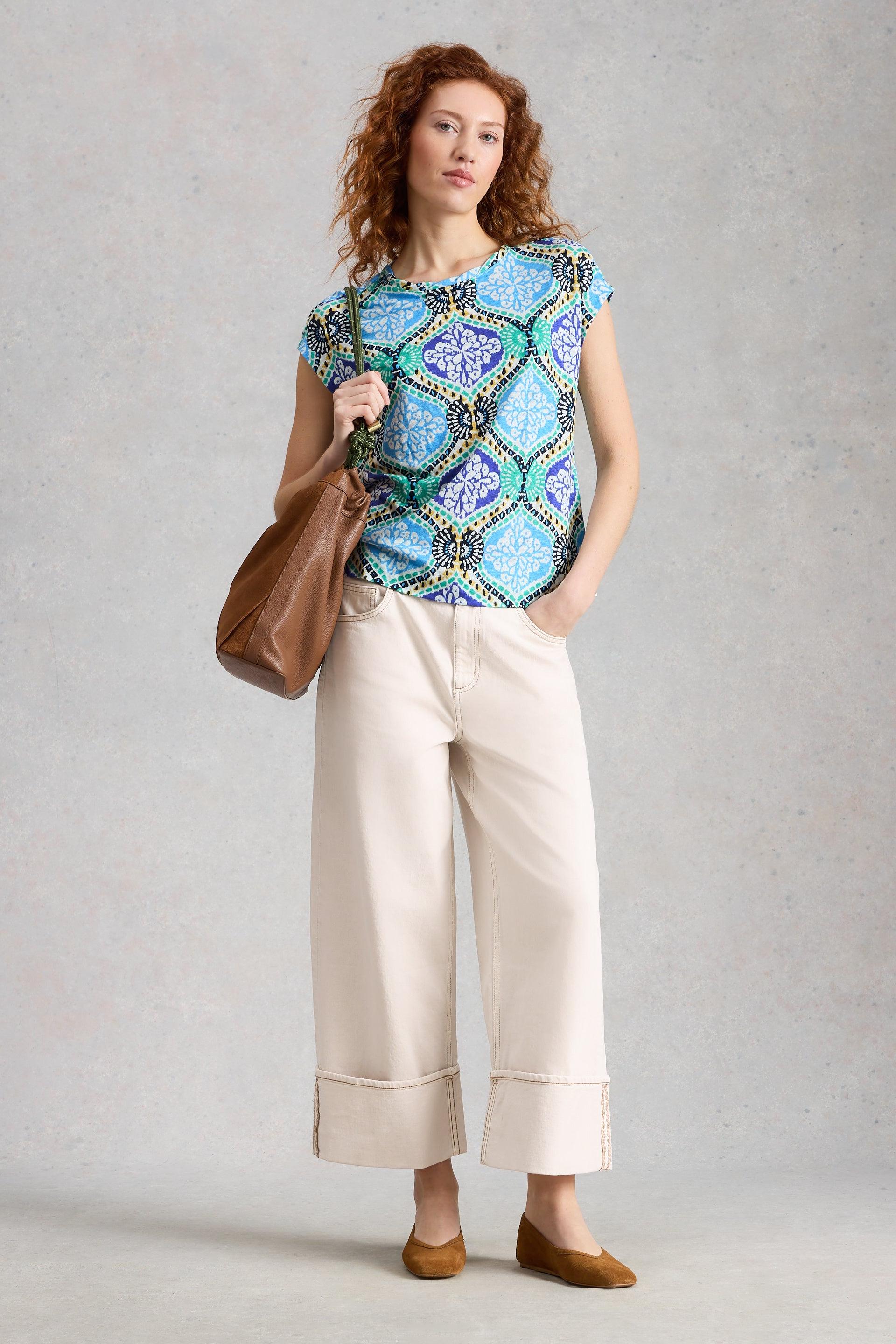 Woman wearing a patterned White Stuff top and beige pants, holding a brown bag on a gray background