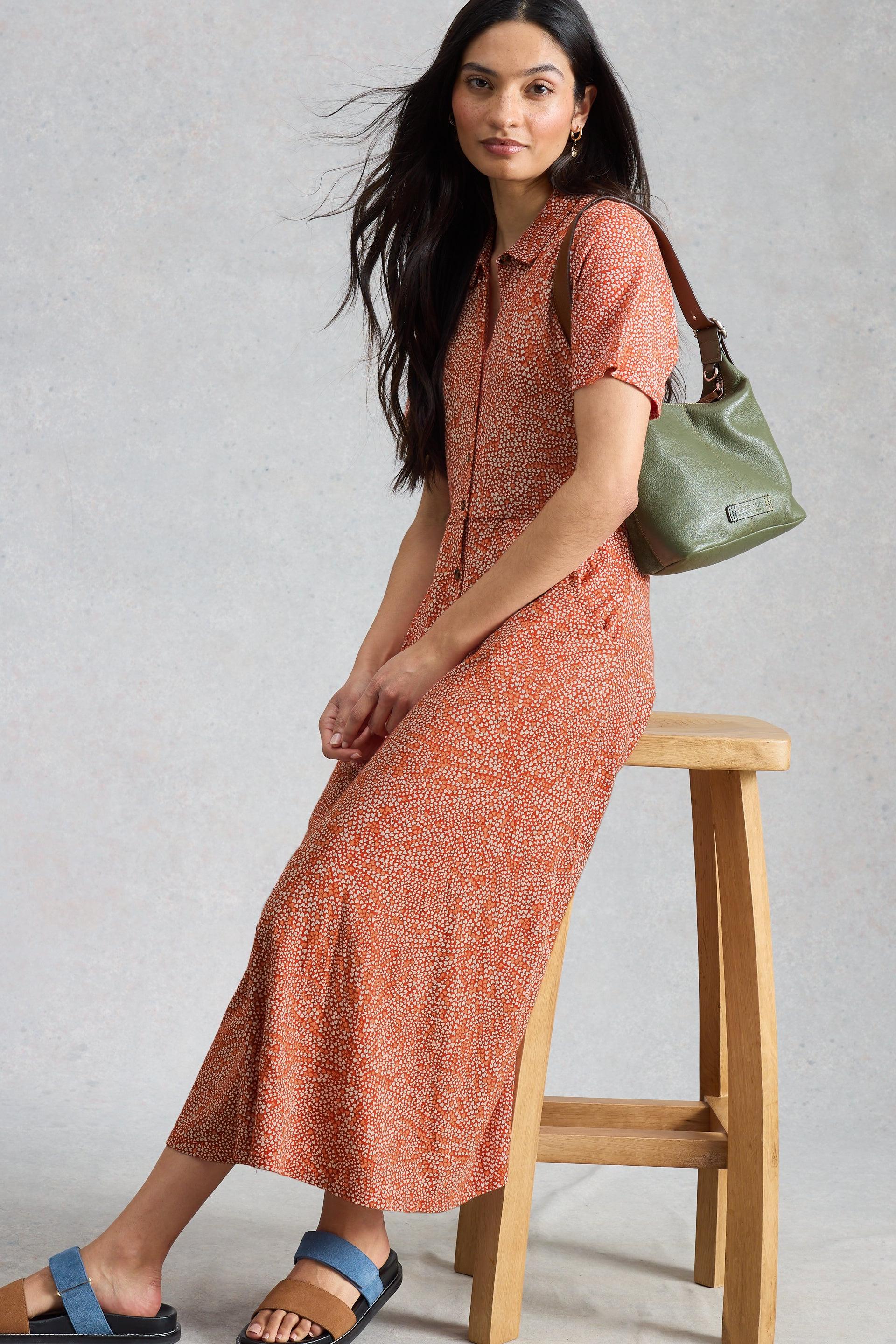 Woman in a patterned organic cotton dress holding a green bag, standing on a wooden stool against a plain background