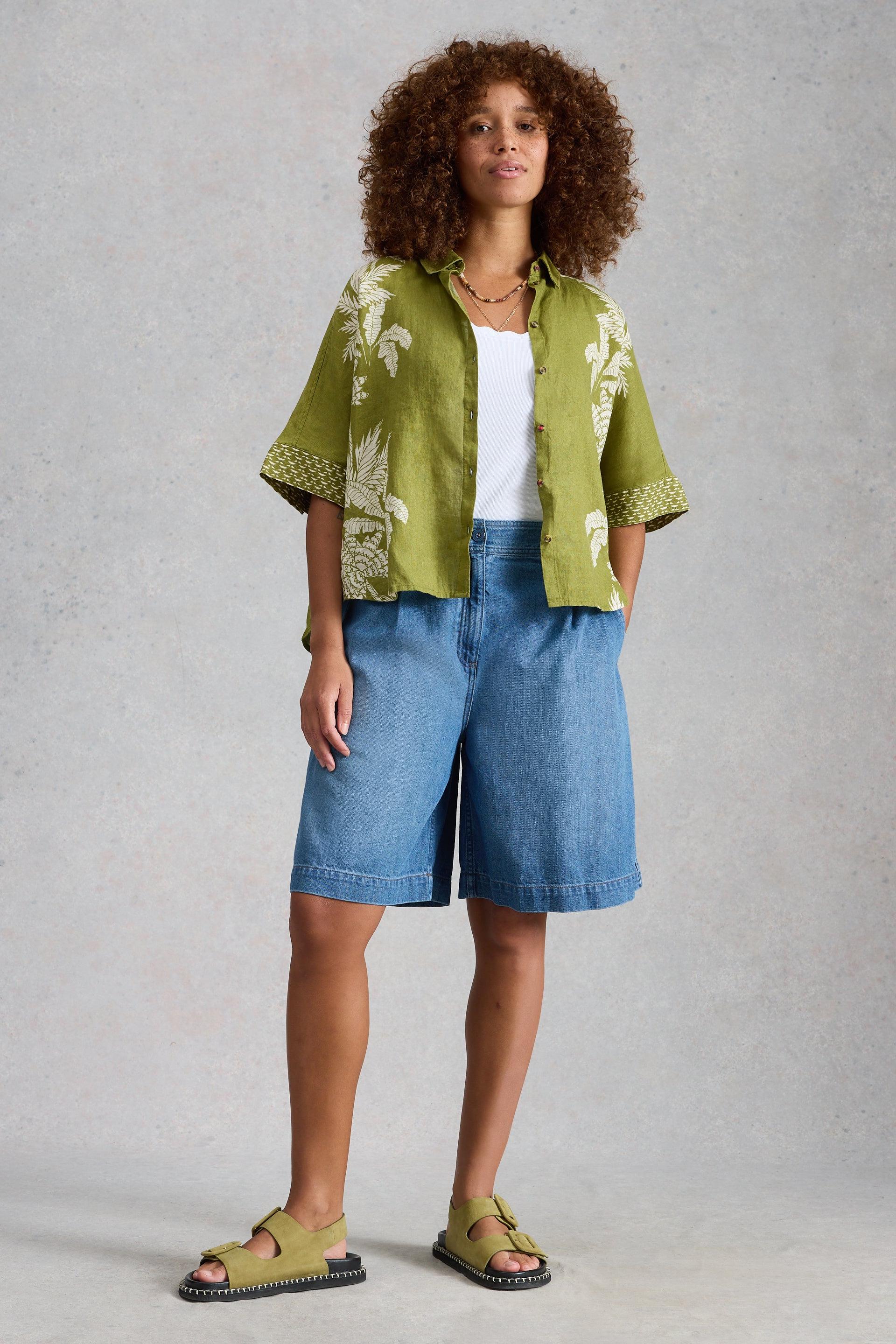 Woman wearing a green floral kimono-style jacket, white top, blue organic cotton denim shorts, and green sandals against a gray background.