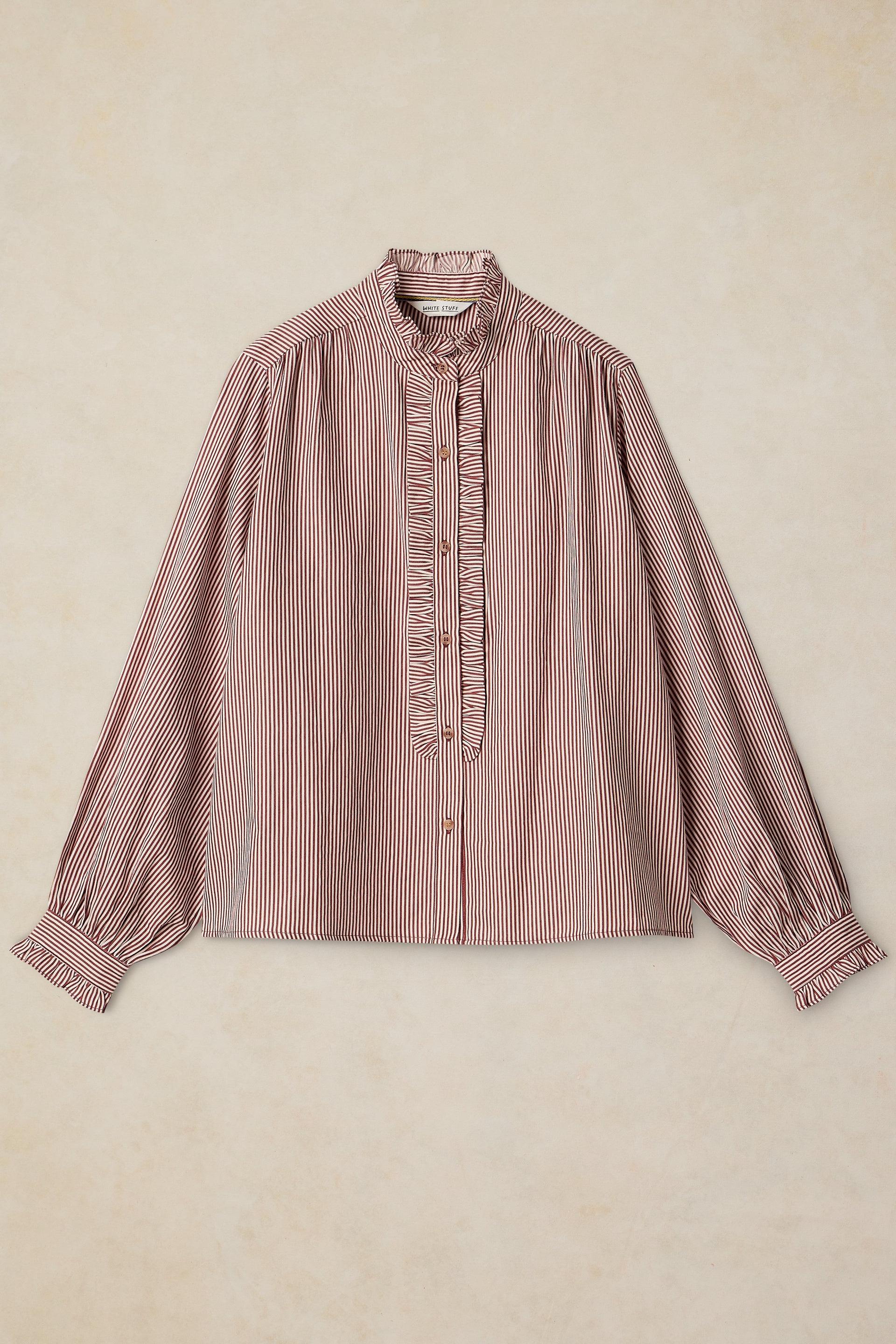 A long-sleeve blouse with vertical stripes in dusty pink and white, featuring a high ruffled collar and a ruffled placket down the front center, laid flat against a textured beige background.
