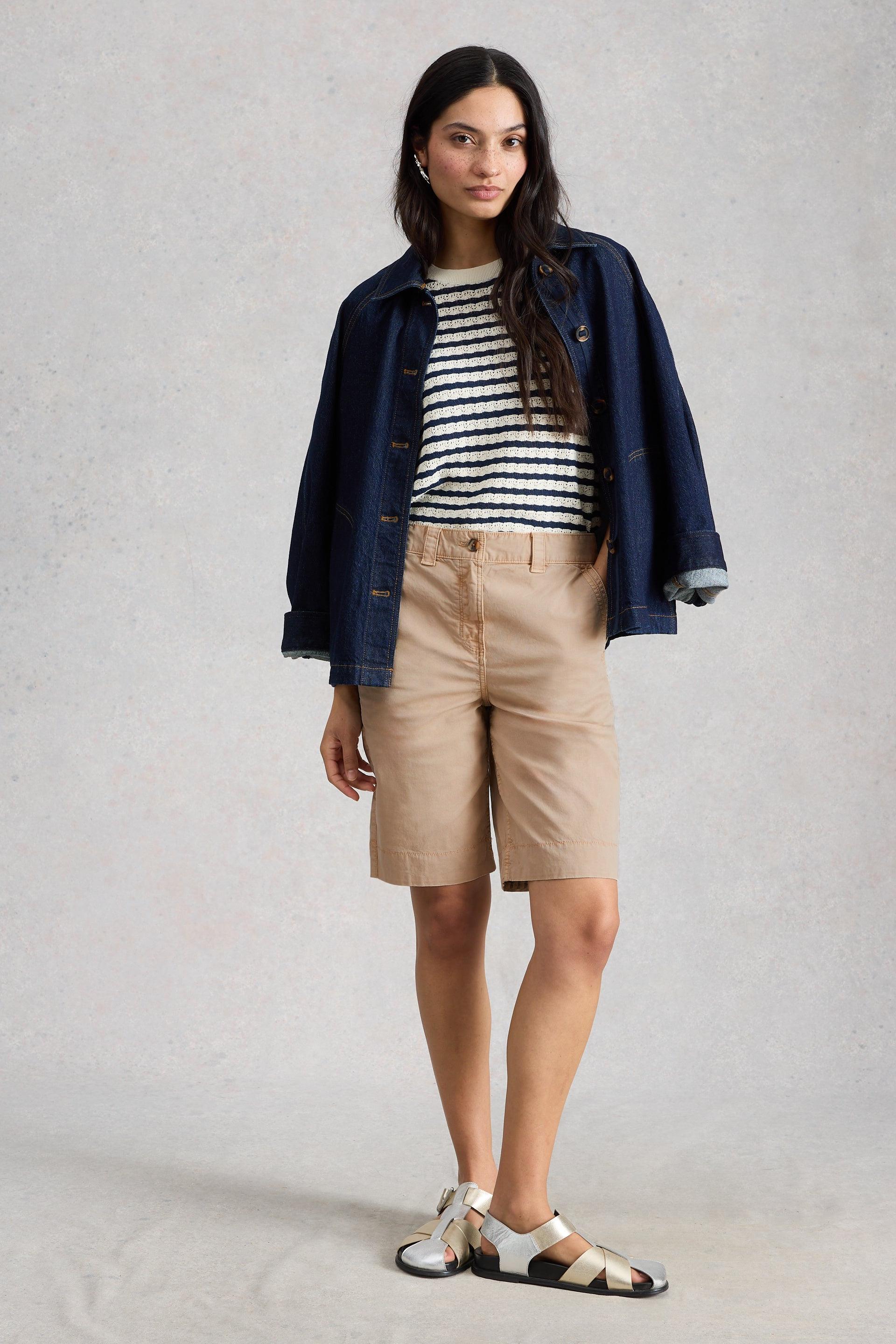 Woman wearing a striped shirt, navy jacket, beige shorts, and sandals on a plain background