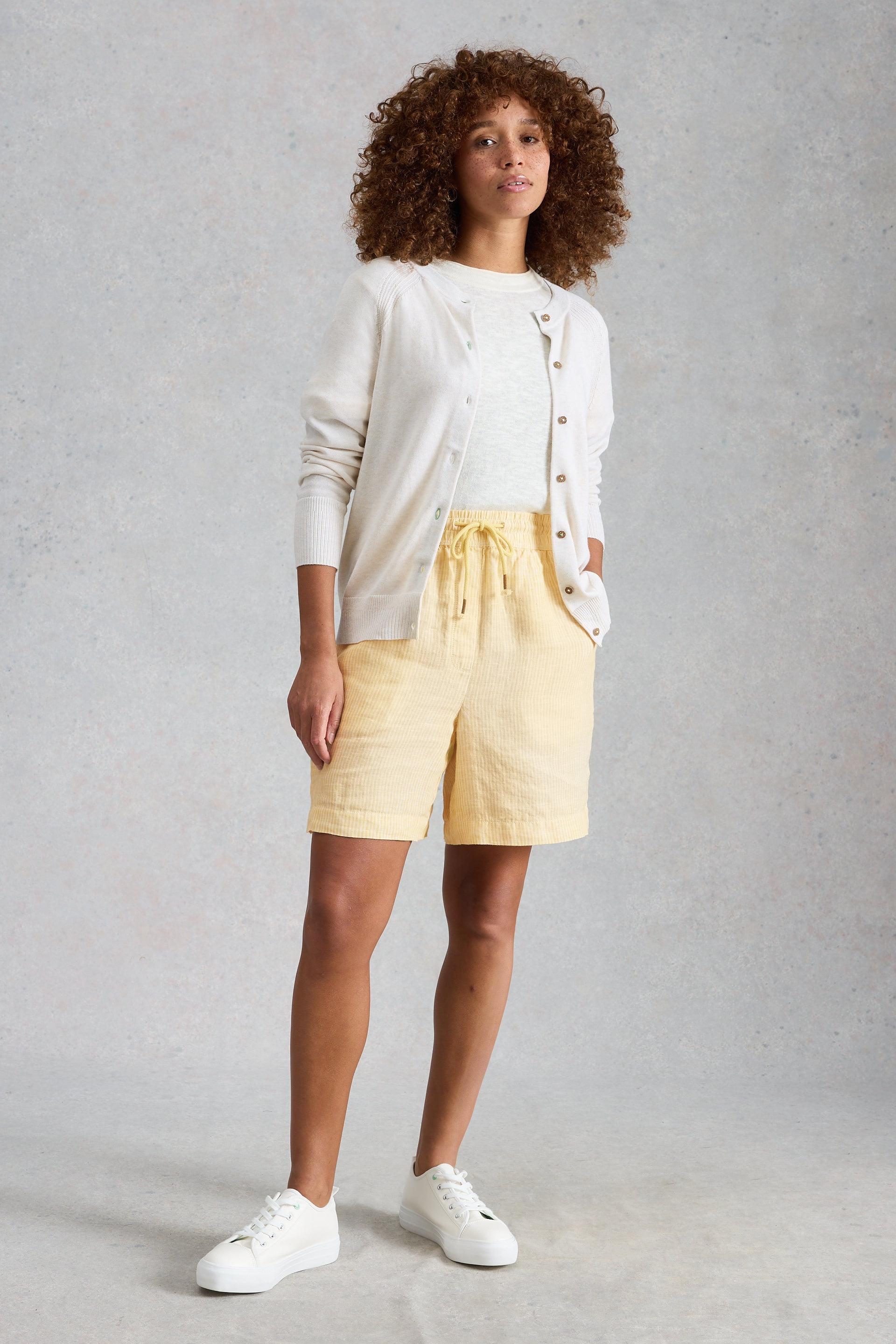 Woman wearing a white cardigan, light yellow linen shorts, and white sneakers on a gray background
