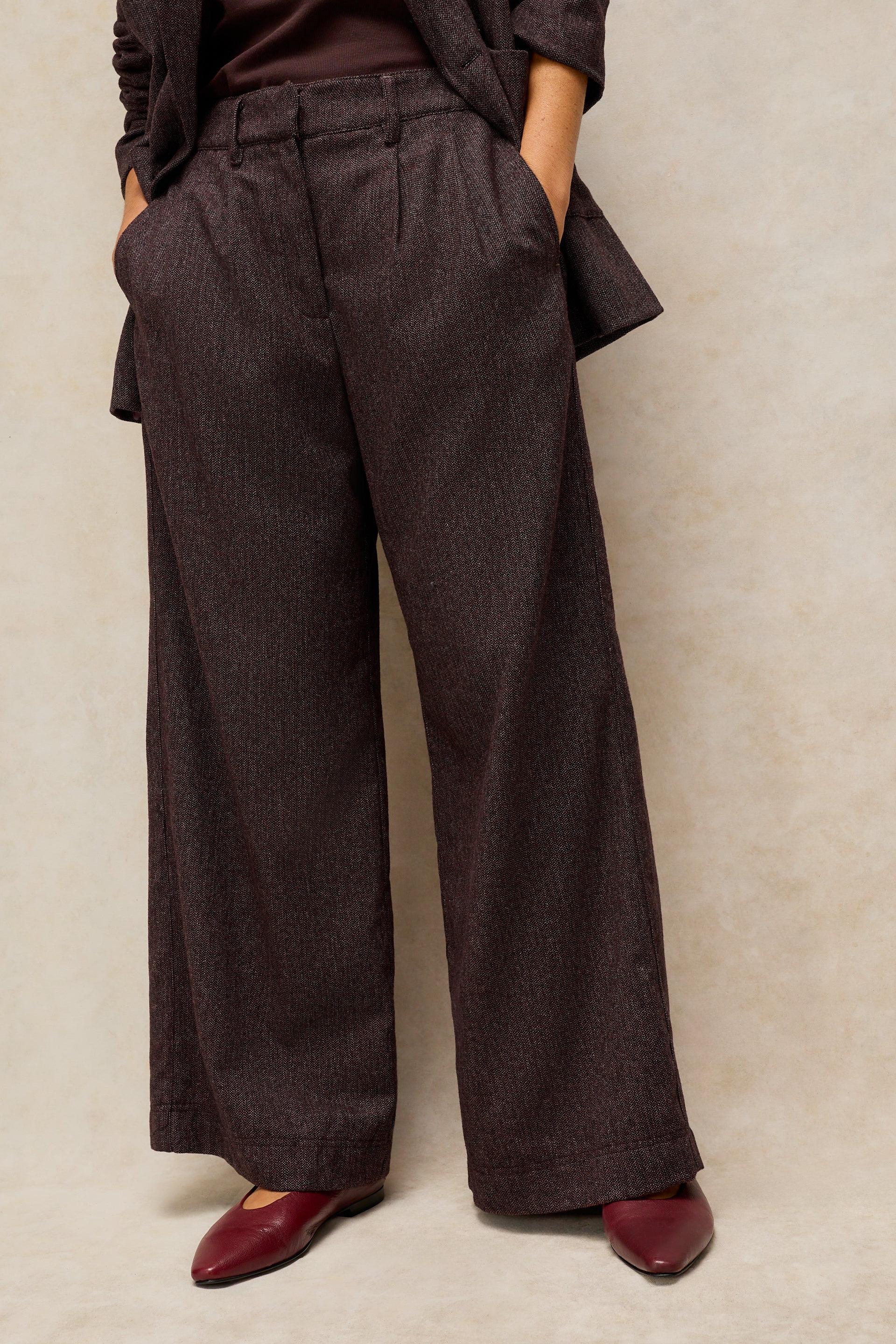 A person wearing dark brown wide-leg trousers with a subtle heathered texture, paired with a dark top and burgundy pointed flats, standing against a textured beige studio backdrop.