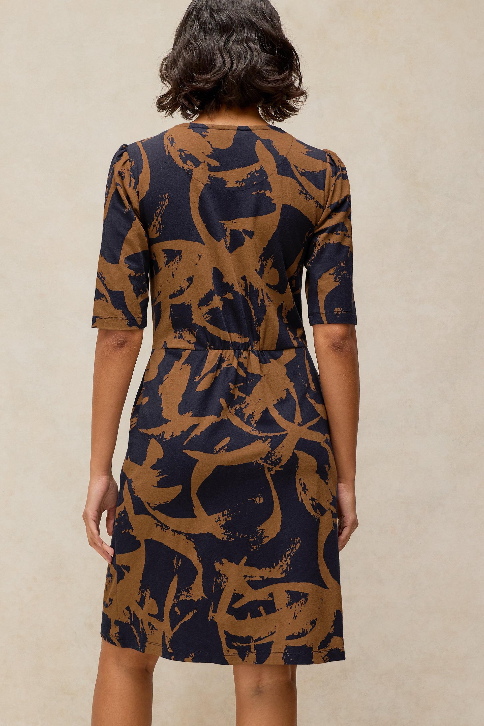 Rear view of a woman wearing a dark brown and black patterned organic cotton dress against a neutral background.